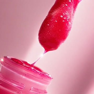 Close-up of a pink lip gloss applicator dripping shiny, glittery pink gloss into an open cosmetic jar.