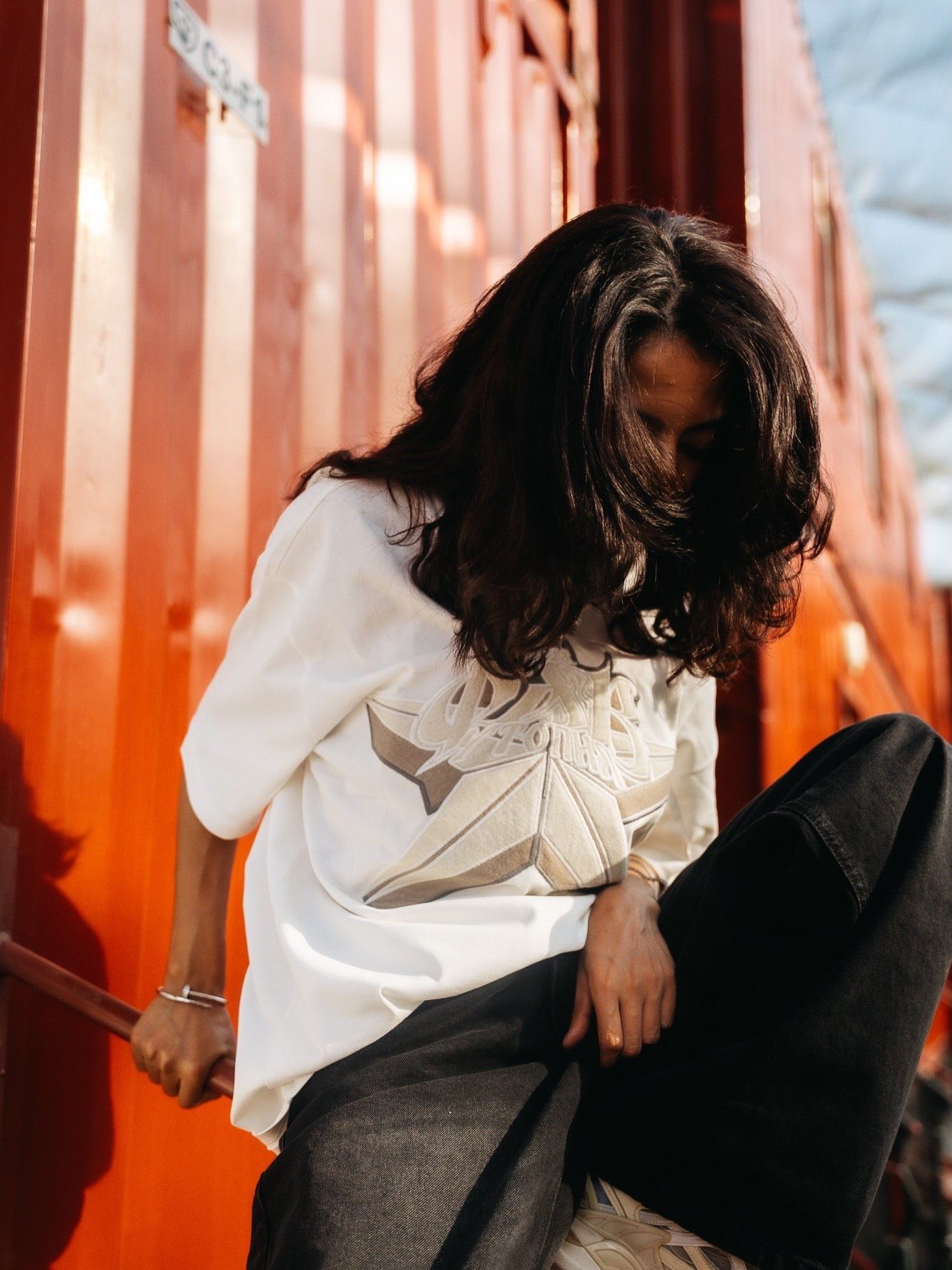 Woman in white blouse and dark jeans posing against a vibrant red shipping container outdoors.
