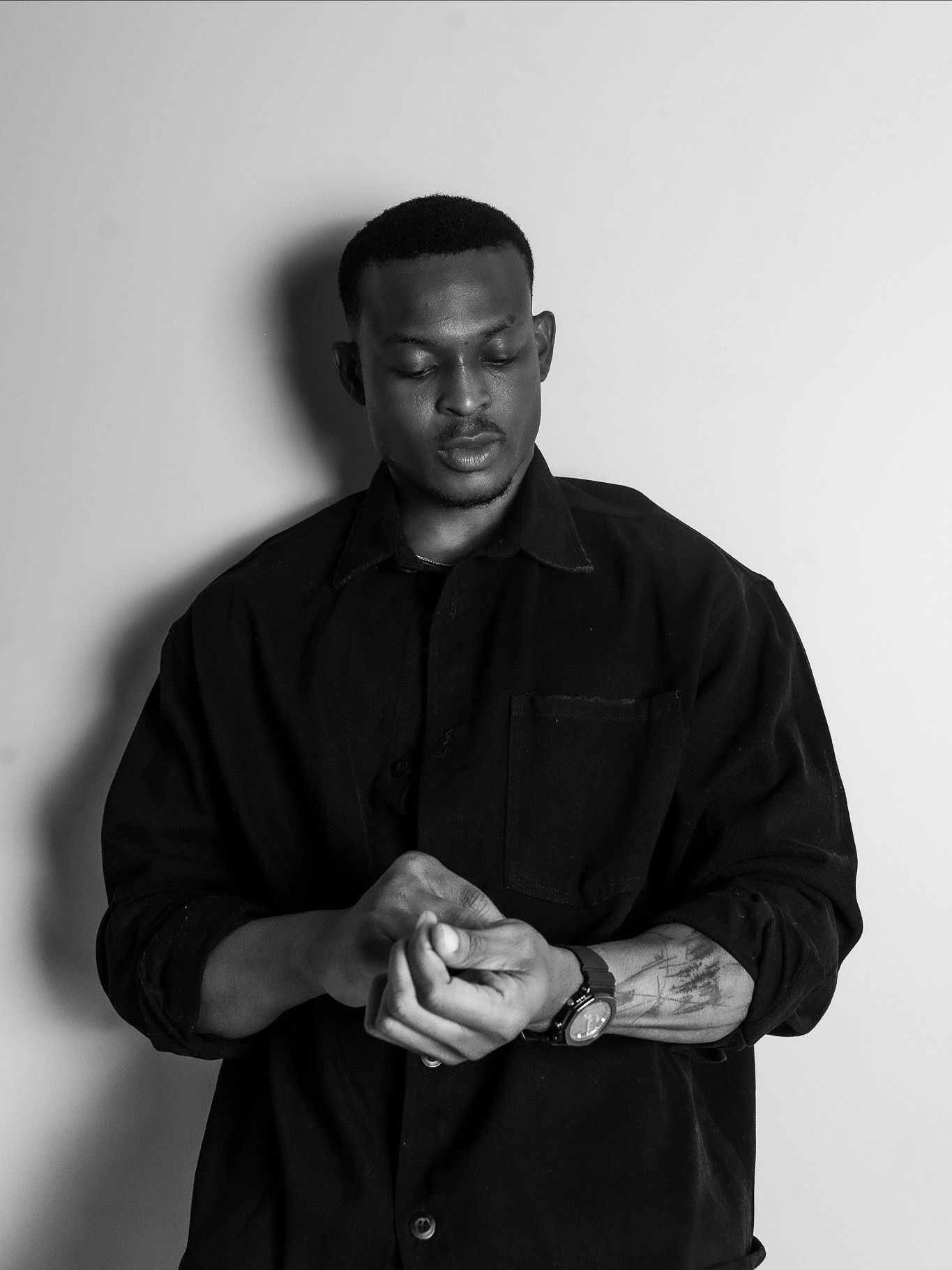 Black and white portrait of a man in a black shirt adjusting his watch, tattooed arm visible.