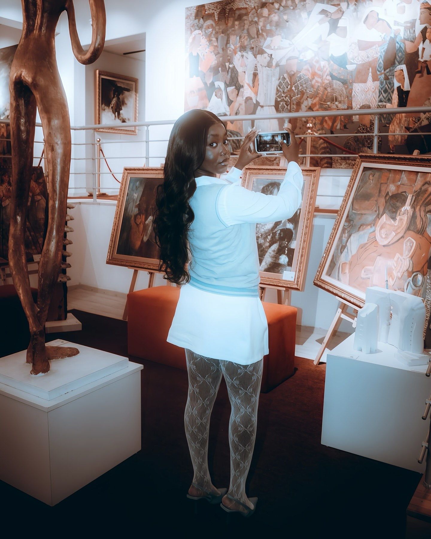 Woman in white outfit photographing artwork in a gallery filled with framed paintings and sculptures.