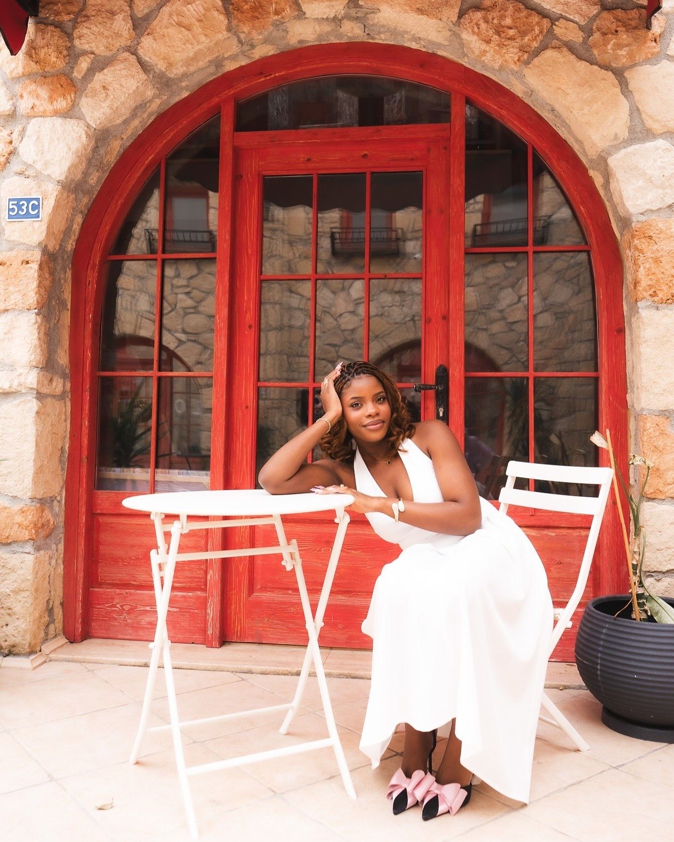Woman in white dress seated at white bistro table before red arched door of stone building.