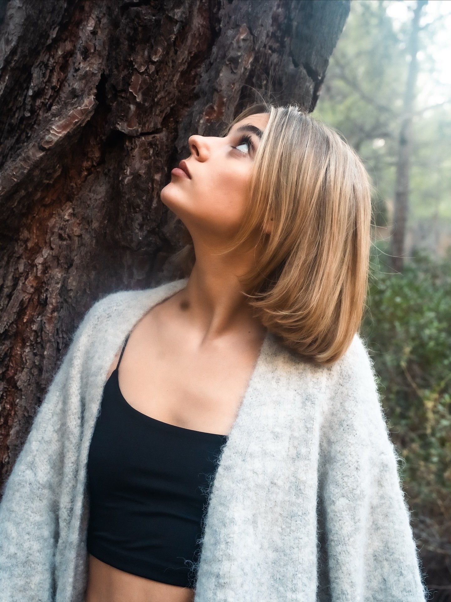 Young blonde woman in gray cardigan and black crop top leans against tree bark, gazing upward outdoors.