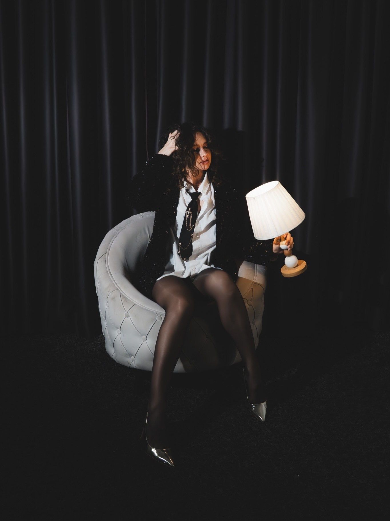 Woman in black blazer and tie sits in white tufted chair holding small lamp in dark dramatic studio.