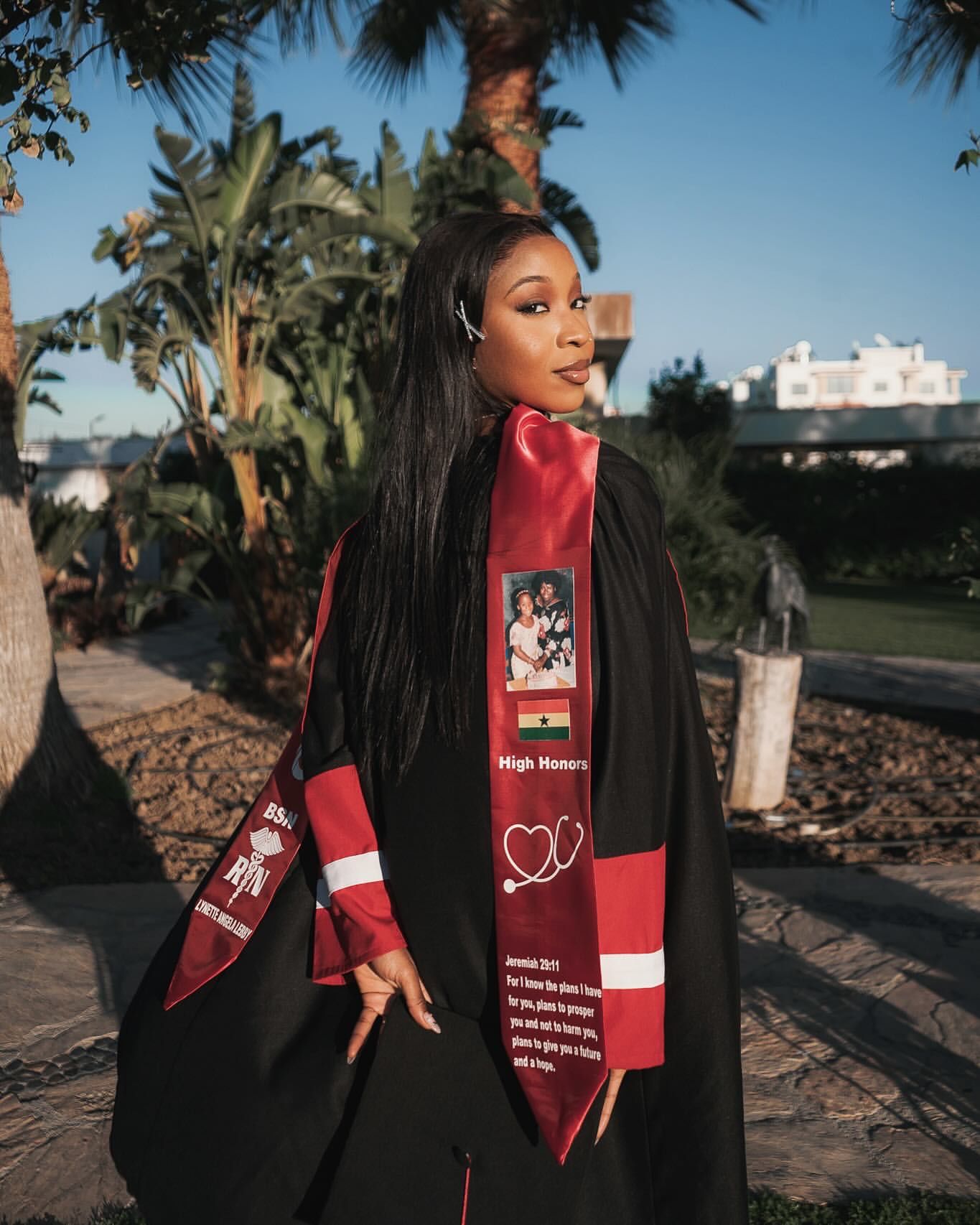 Black woman in graduation regalia with High Honors stole poses outdoors at golden hour.