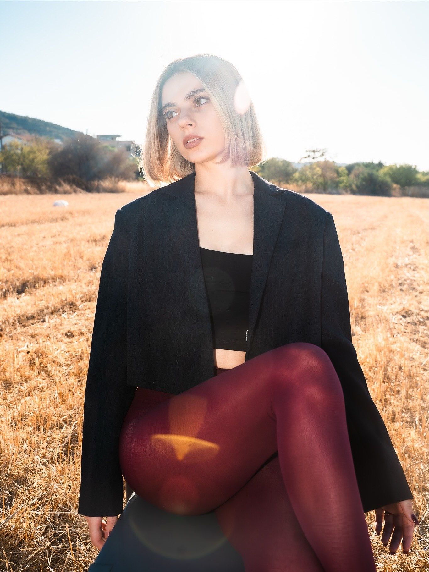 Woman in black blazer and burgundy tights sitting in a sunlit golden field, posing with knees up.