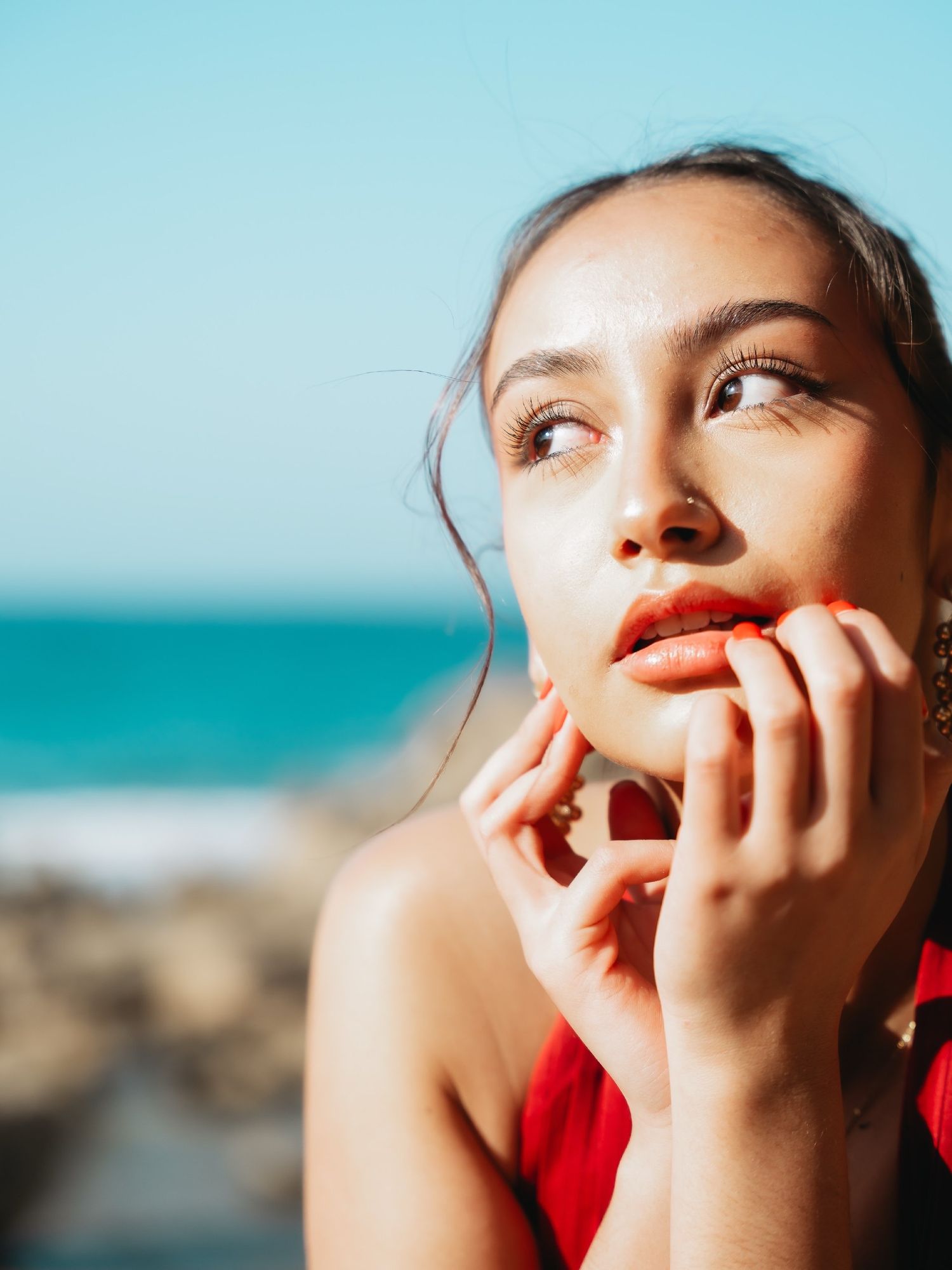 Young woman in red top gazes upward at beach, hands near face, with turquoise ocean in background.