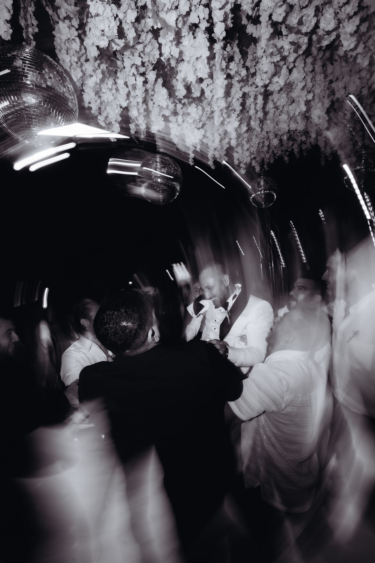 Black and white photo of elegantly dressed guests dancing at a wedding reception under floral ceiling decor.