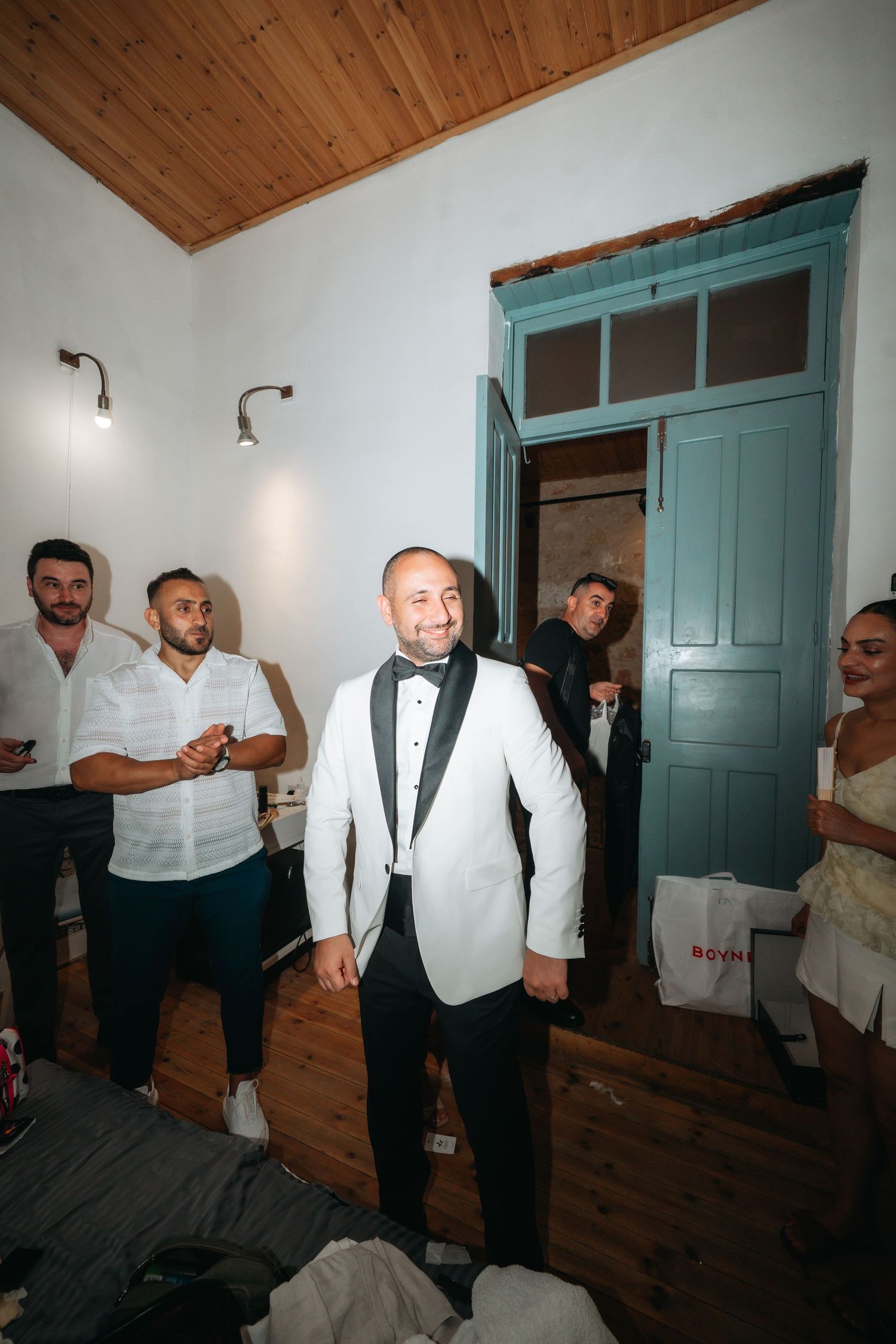 Groom in white tuxedo jacket smiles with groomsmen in decorated room with teal door.