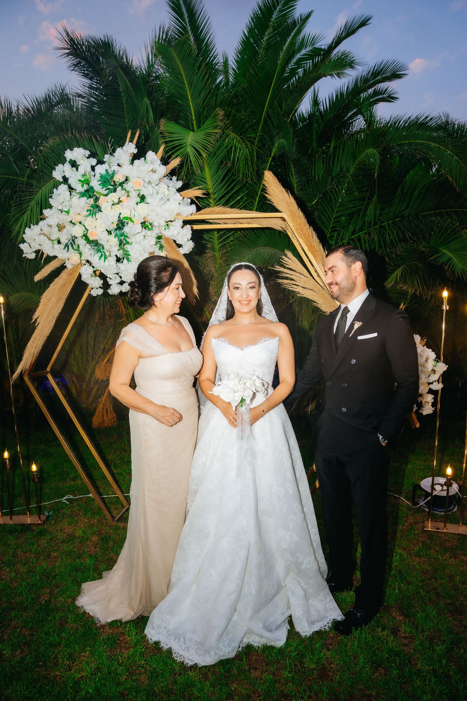 Bride in white gown holds bouquet beside groom in black suit and woman in beige dress at outdoor wedding ceremony.