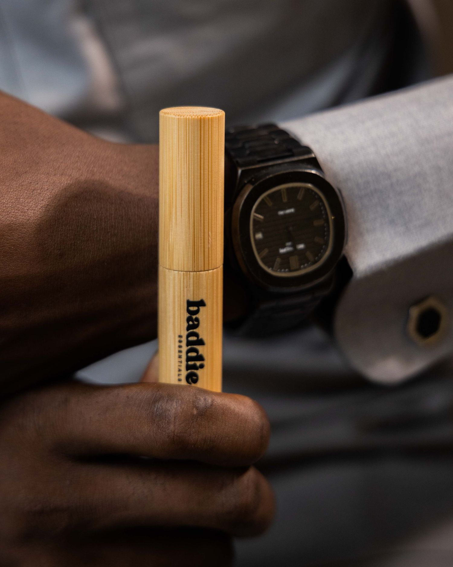 Hand holding a bamboo Baddies cosmetic tube, wearing a black square watch with gray sleeve visible.