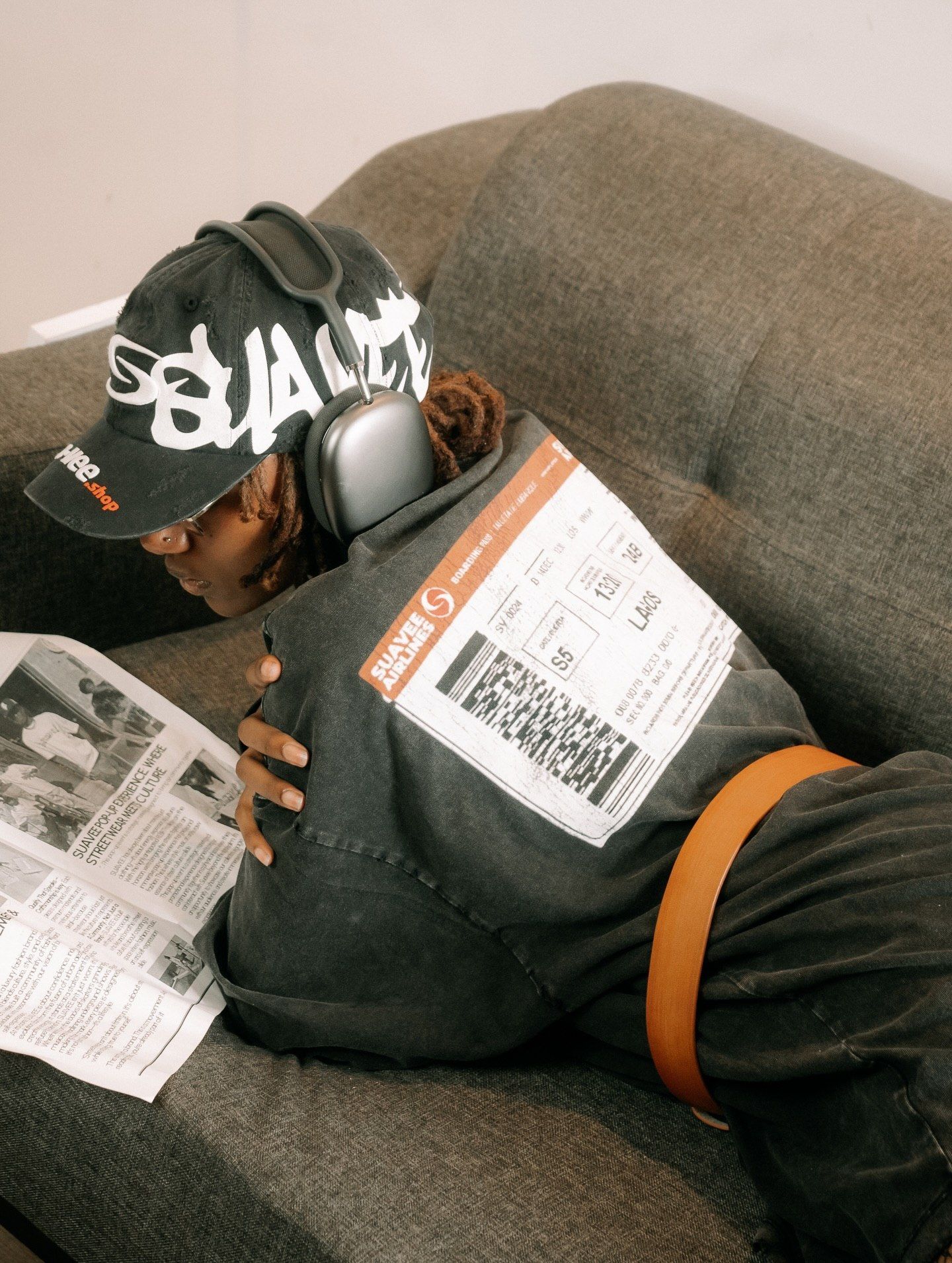 Person in a cap and headphones relaxing on a gray sofa, holding a newspaper, wearing a dark hoodie with a shipping label.
