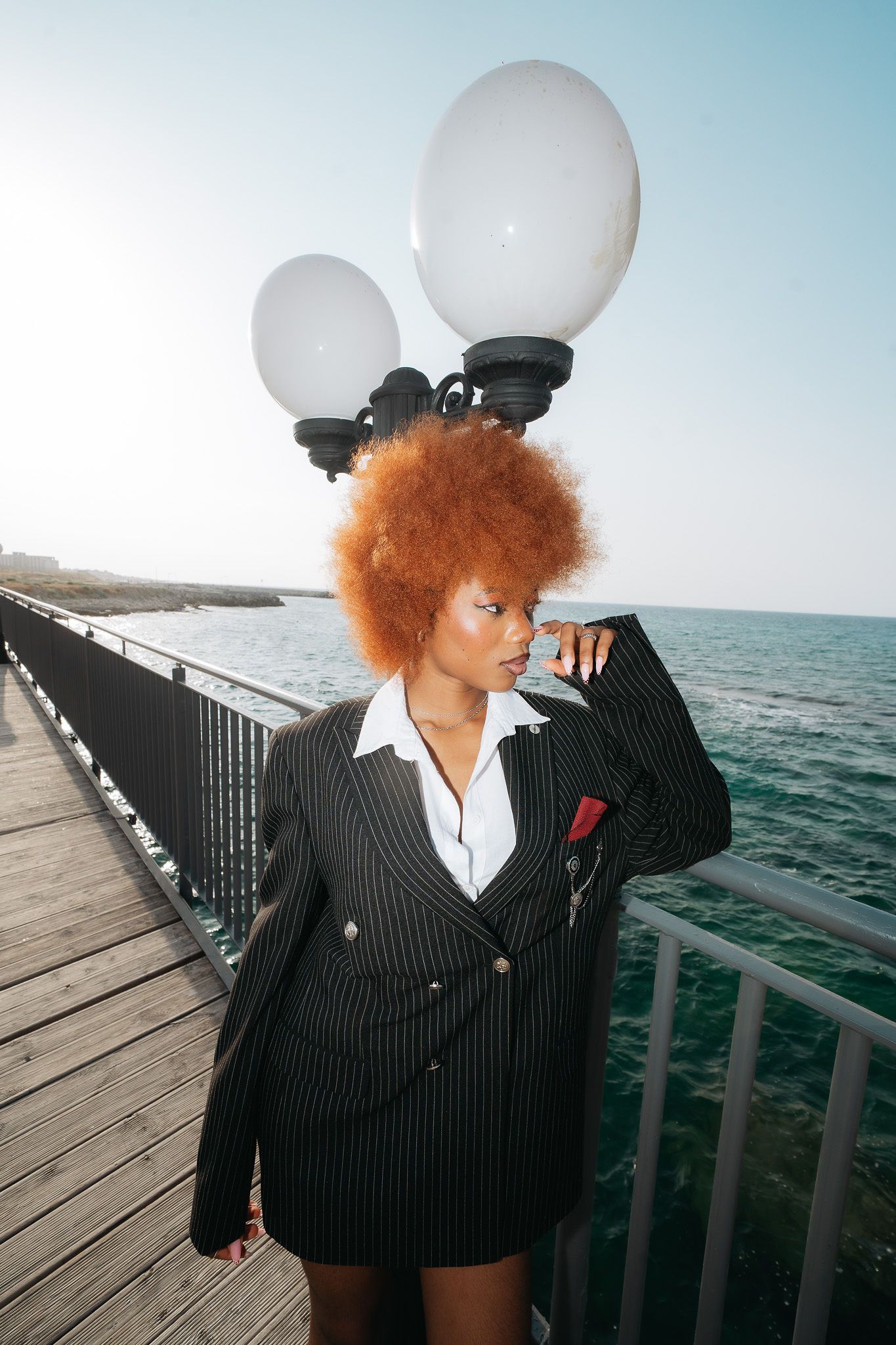 Woman with auburn afro in pinstripe blazer poses by seaside lamp post on wooden pier.