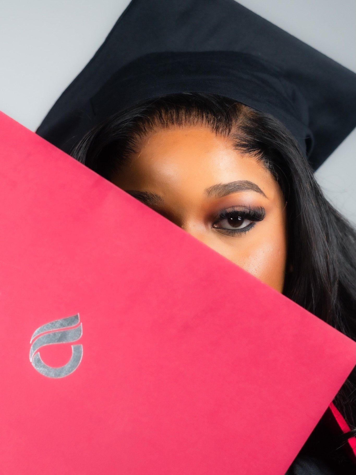 Graduate peeking over red diploma folder wearing black mortarboard cap.