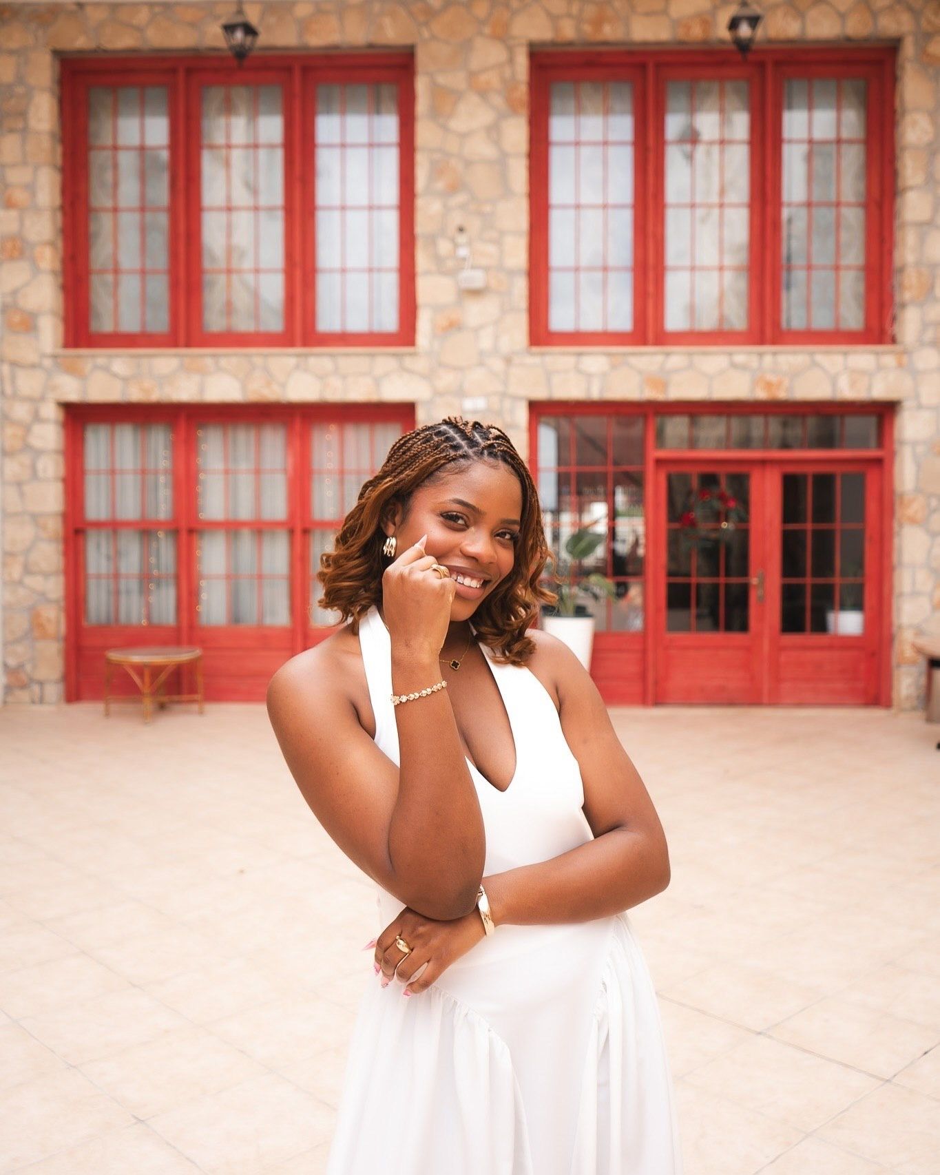 Young woman in white halter dress smiling outdoors near stone building with red windows and doors.