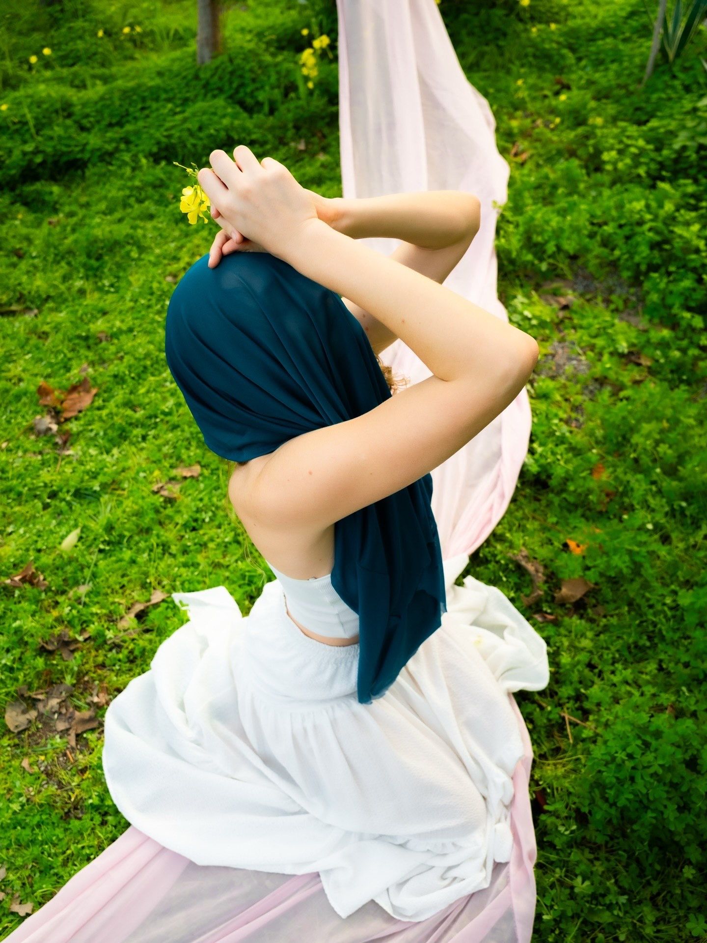 Woman in white dress with teal hair wrap holding yellow flower, seated outdoors on lush green grass.
