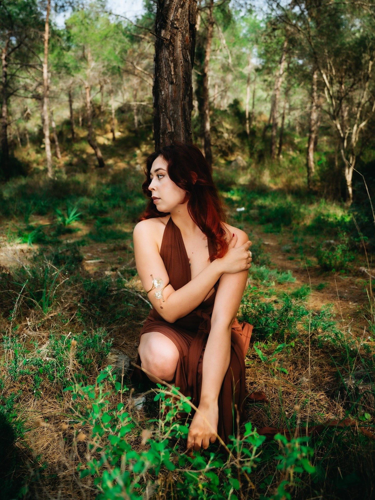 A young woman in a brown dress sits thoughtfully in a lush forest, hugging her knees beside a tree.
