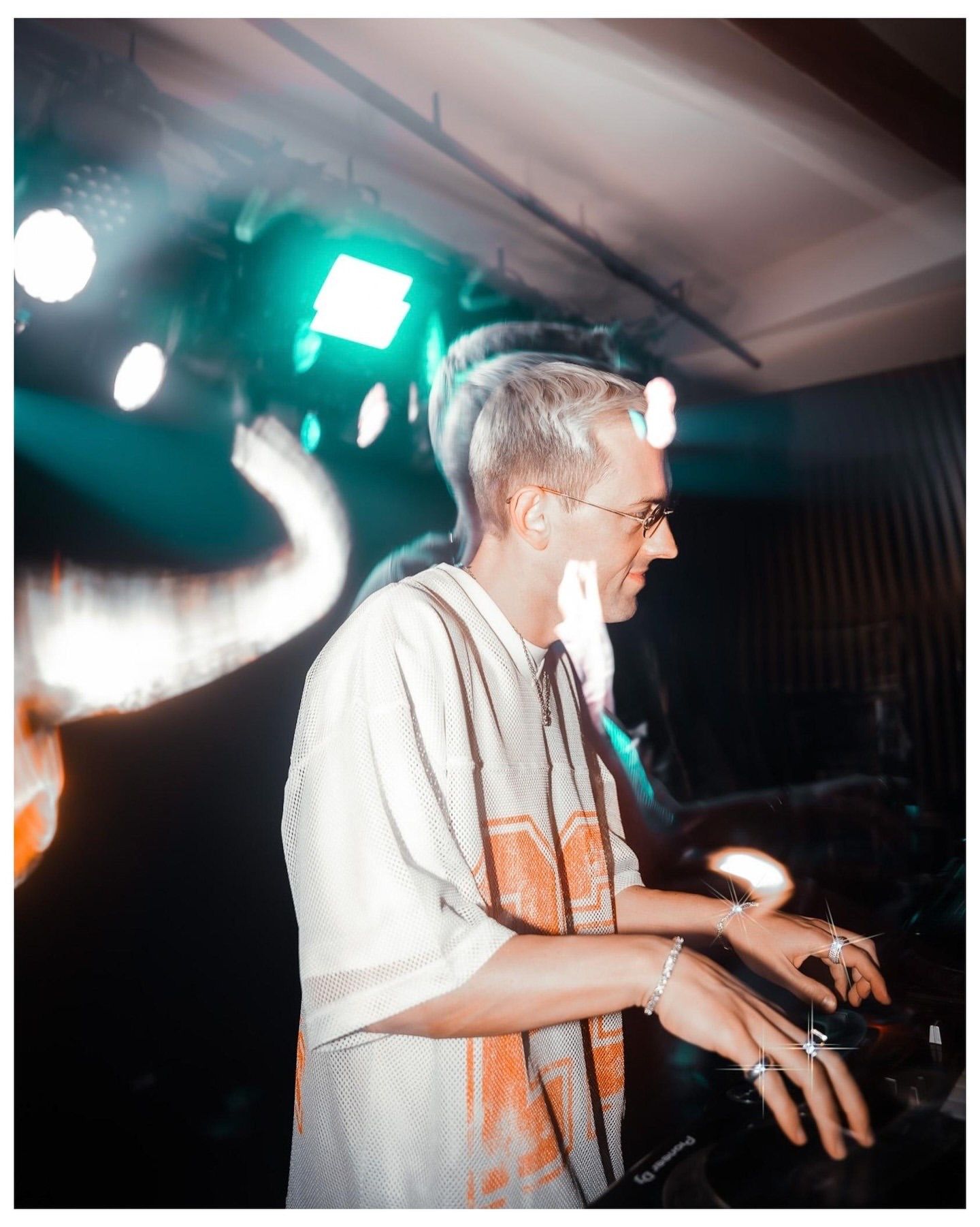 A DJ with white hair and glasses wearing a white shirt performs at a dimly lit venue with colorful stage lighting.