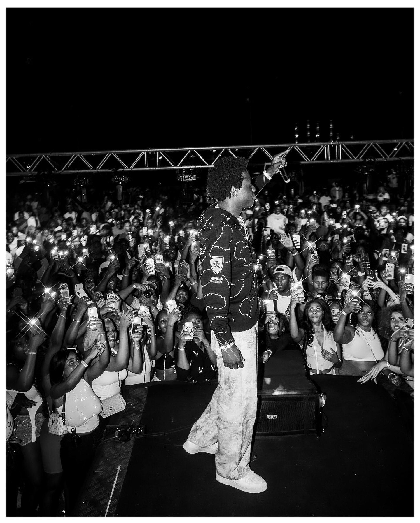 Black and white photo of a performer on stage facing a large crowd holding up phones at a night concert.