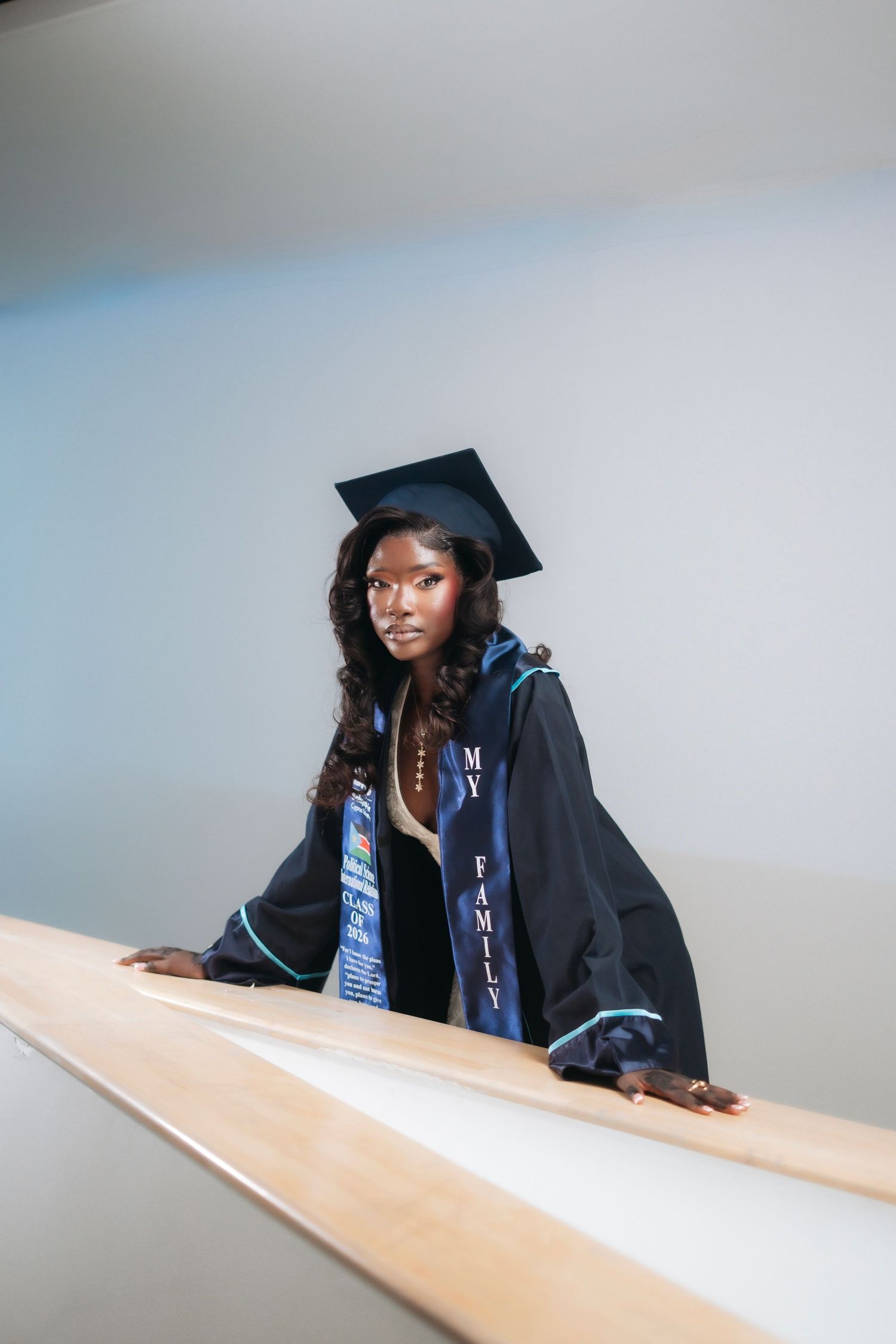 Graduate in cap and gown with 'My Family' stole leans on a railing indoors.