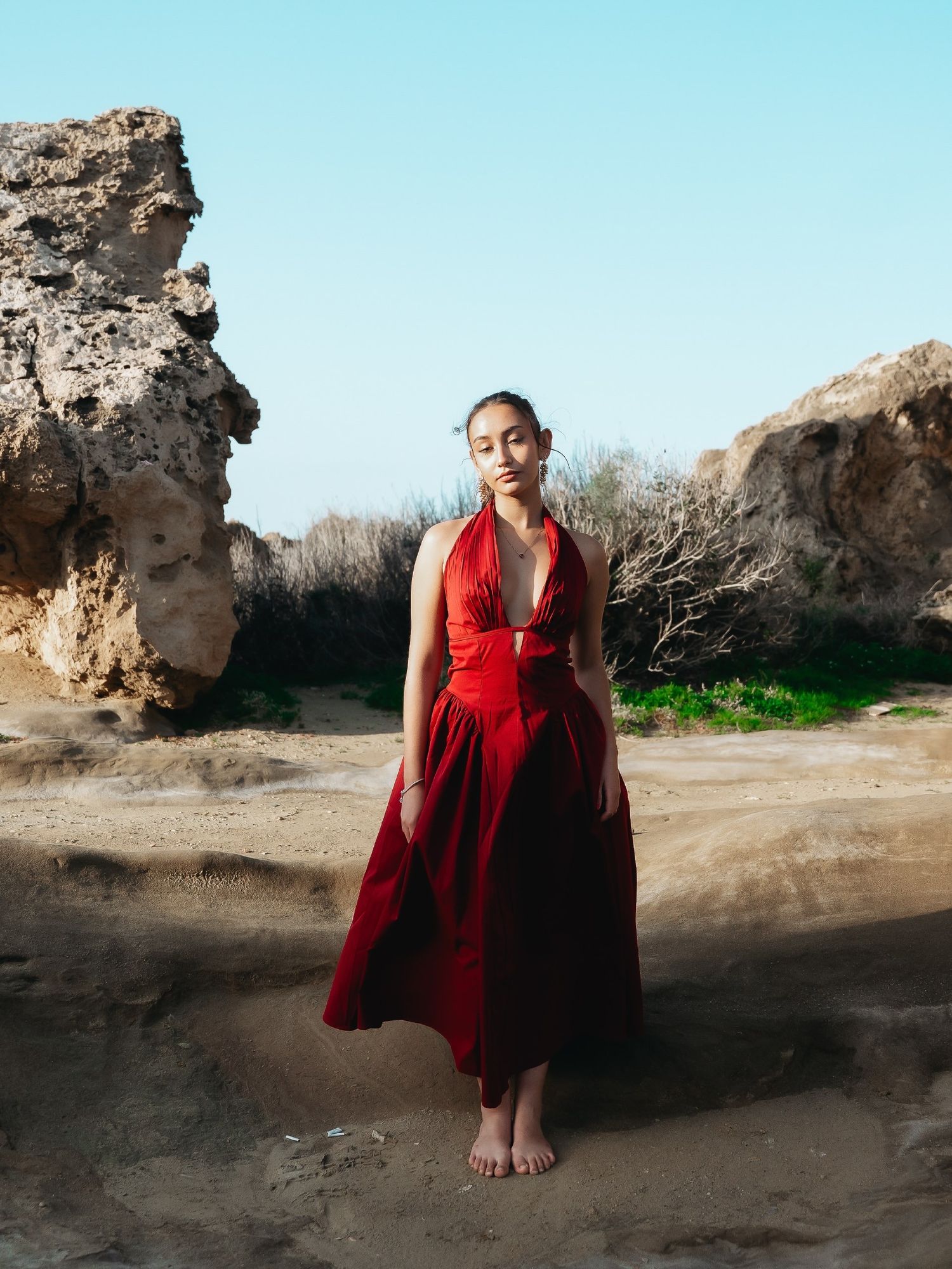 Woman in flowing red halter dress standing barefoot on sandy terrain between rocky formations under blue sky.