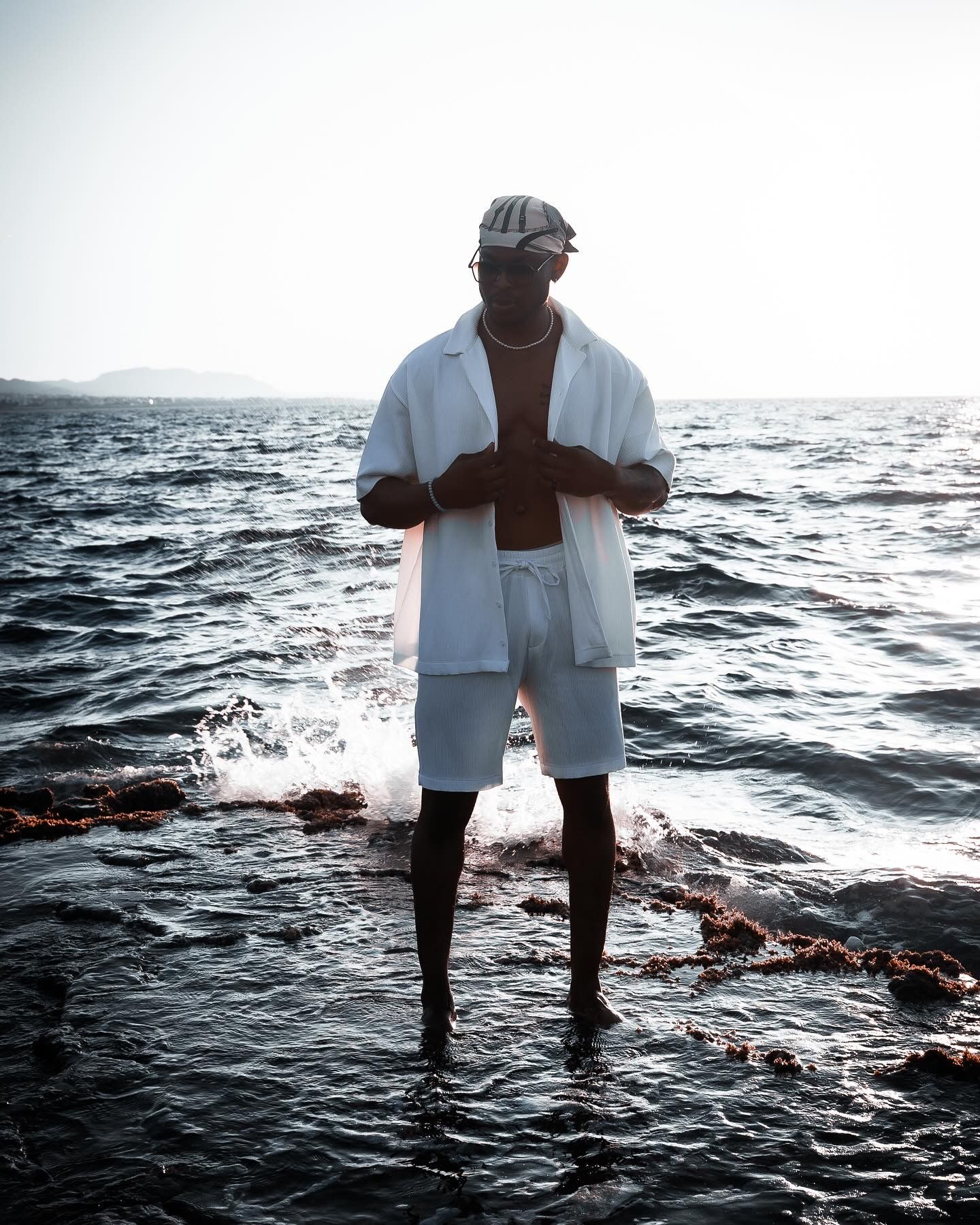 Man in white shirt and shorts standing barefoot on rocky shore with ocean waves and bright sky behind him.