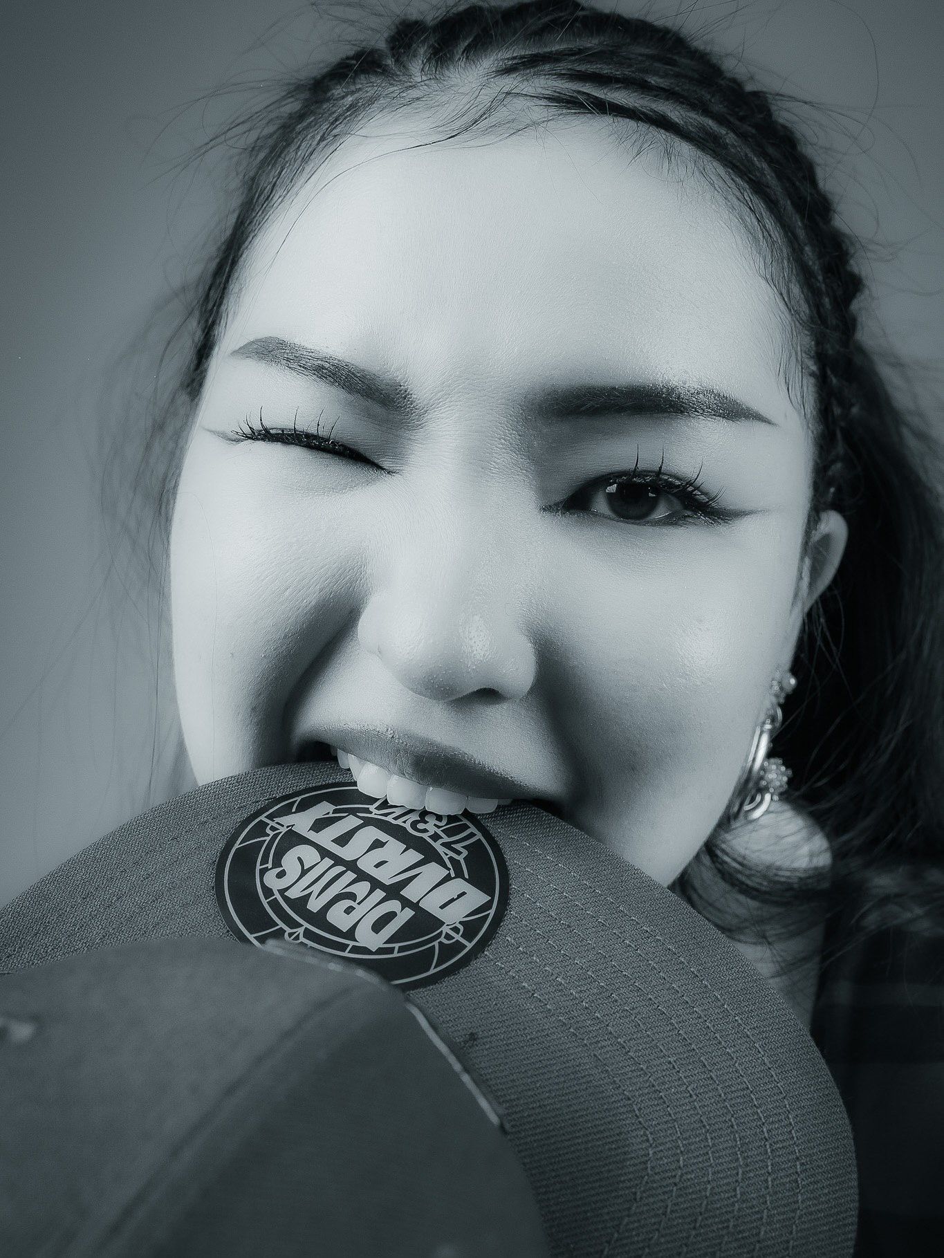Black and white portrait of a young woman biting the brim of a snapback cap, wearing eyeliner and an earring.
