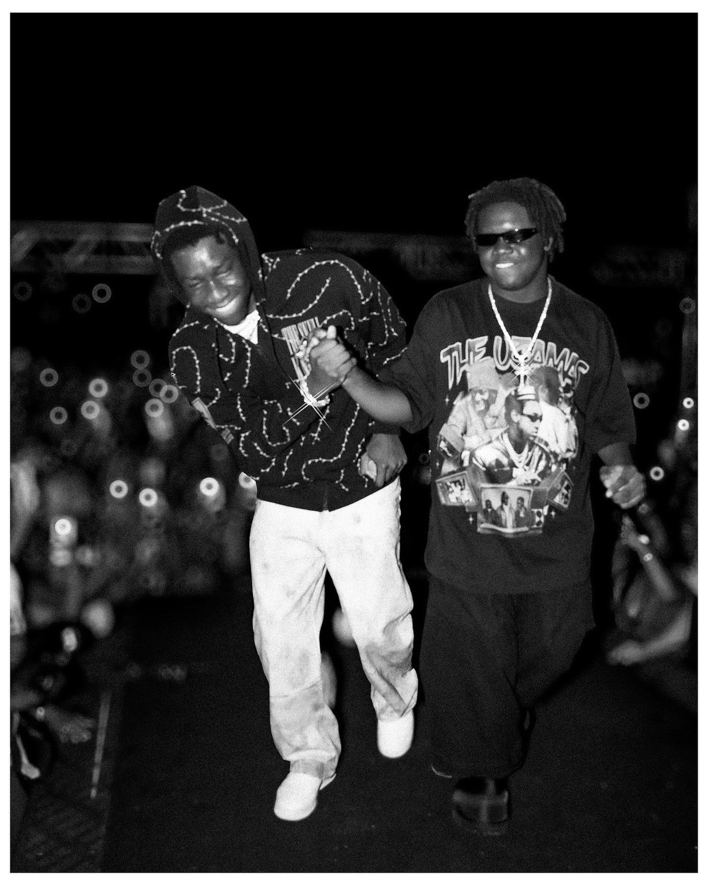 Two men walking on stage at a concert, one in a patterned jacket and white pants, the other in a graphic tee.