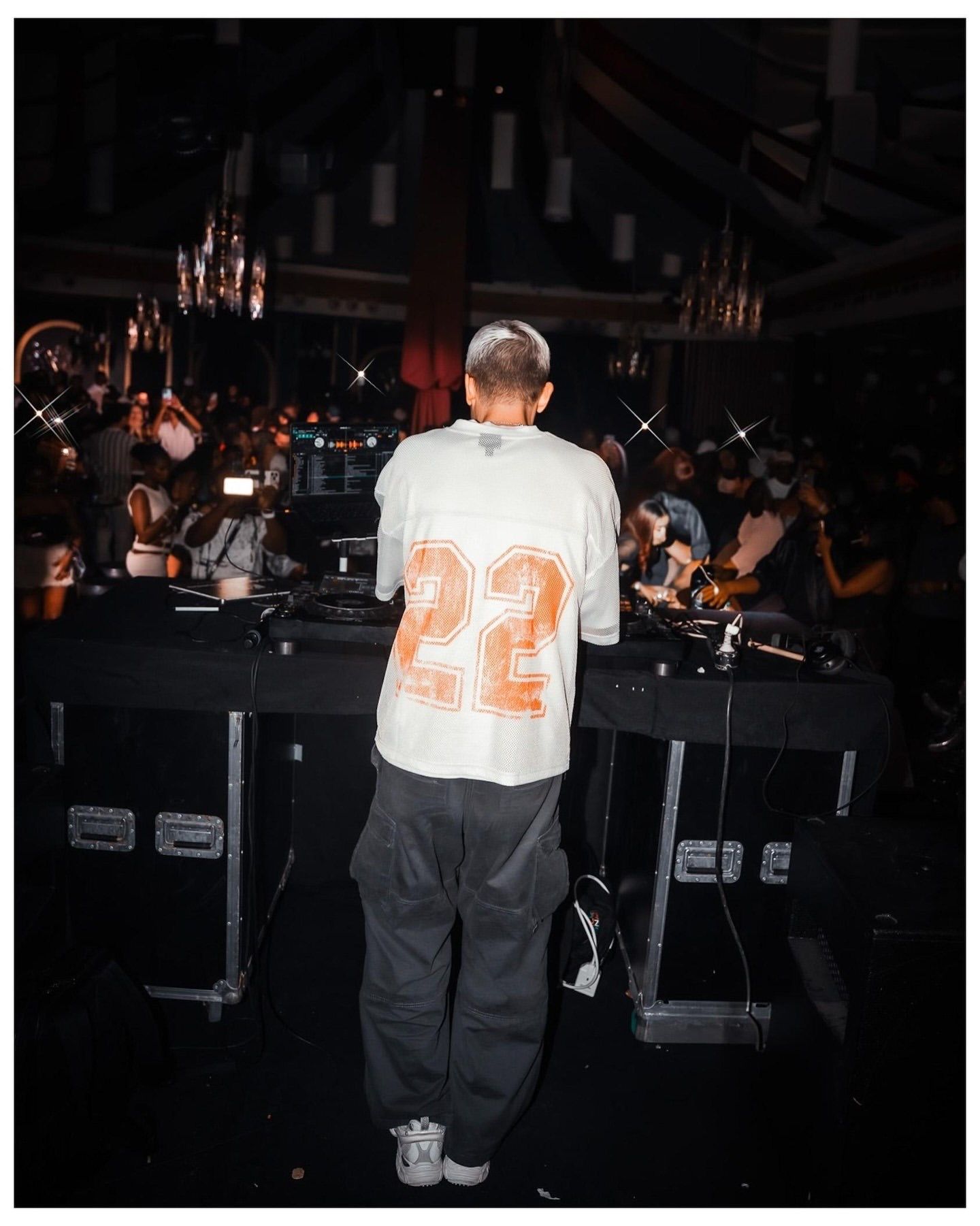 DJ in white jersey number 22 stands at turntables performing for a crowd in a dimly lit venue.