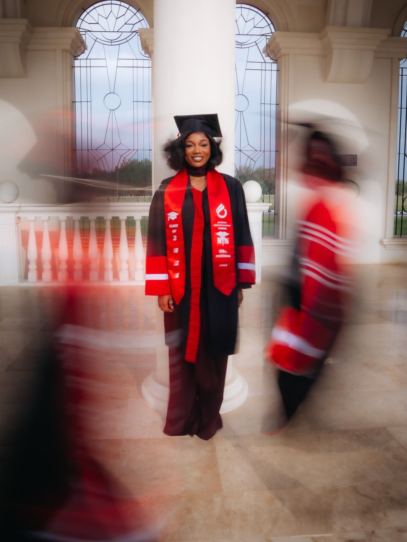 Graduate in black cap and gown with red stole stands among blurred fellow graduates.