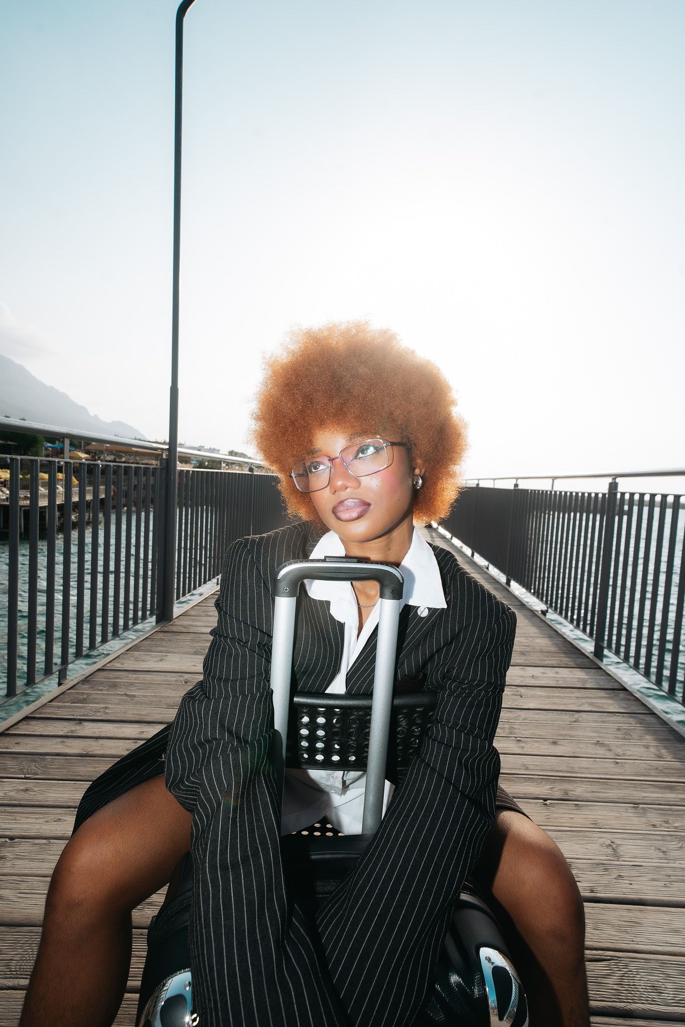 Woman with orange afro in pinstripe suit sits with luggage on sunny waterfront pier.