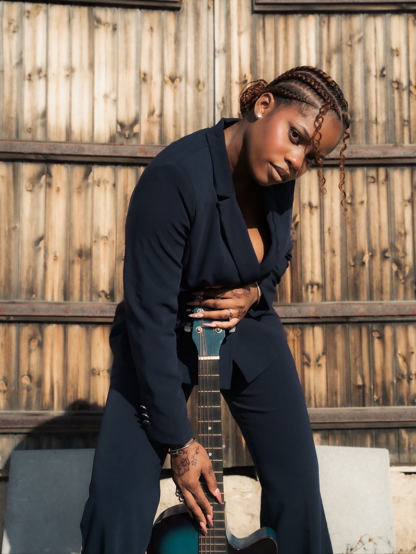 Woman in navy blazer leans against teal acoustic guitar beside wooden fence in moody portrait.