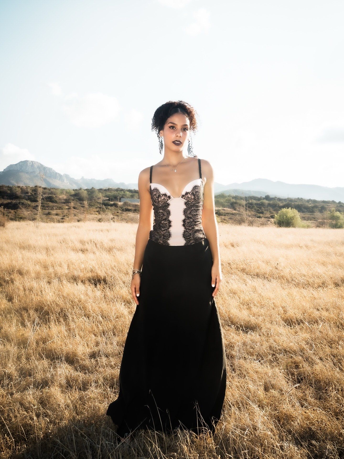 Woman in black and white lace gown standing in golden field with mountain backdrop.