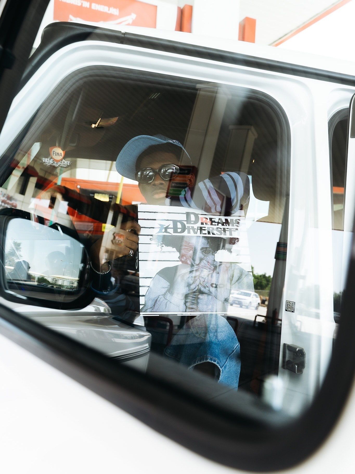 Person holding a Dreams & Diversity sign reflected in a white vehicle's side mirror at a drive-through.
