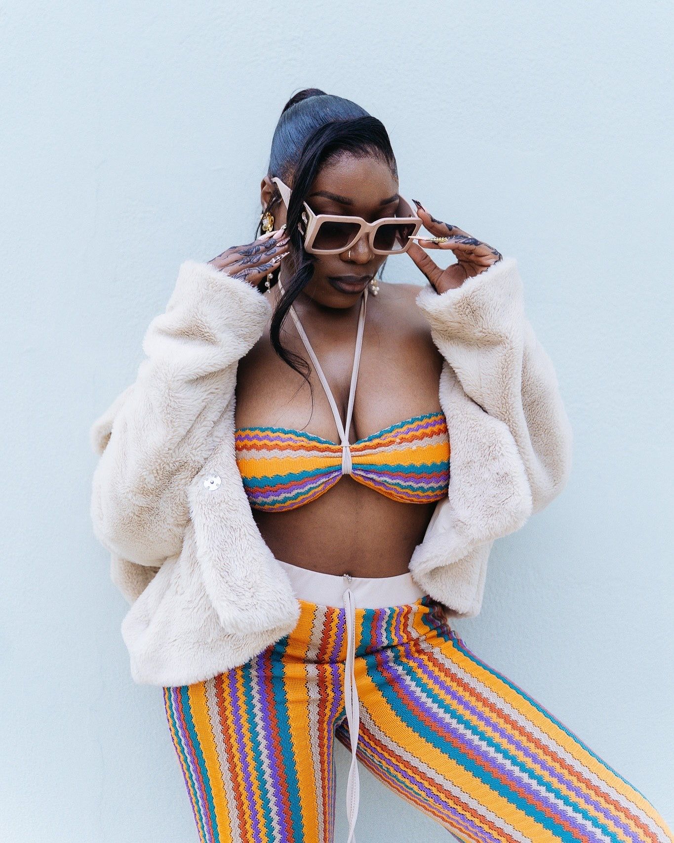 Woman in colorful striped bikini top and pants, fuzzy white jacket, and white sunglasses posing against a light blue wall.