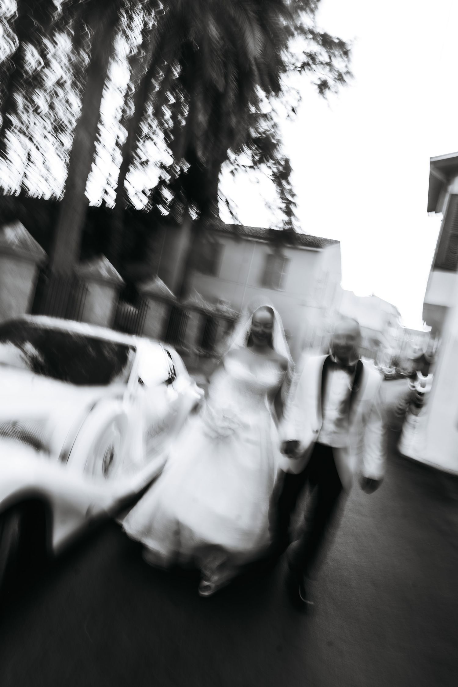 Blurred black and white photo of a bride and groom walking outdoors near a white car on their wedding day.