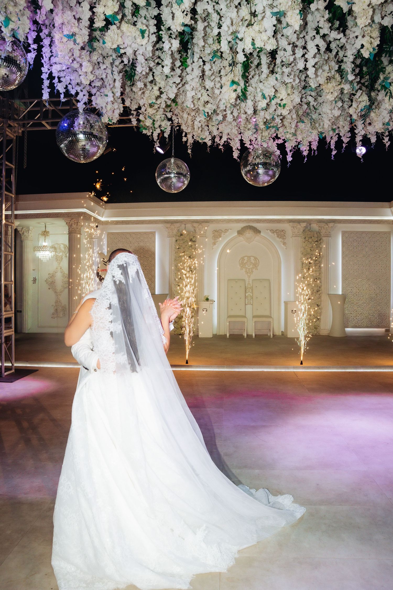 Bride in white gown and veil dancing at elegant wedding reception with floral ceiling and sparkling fountains.