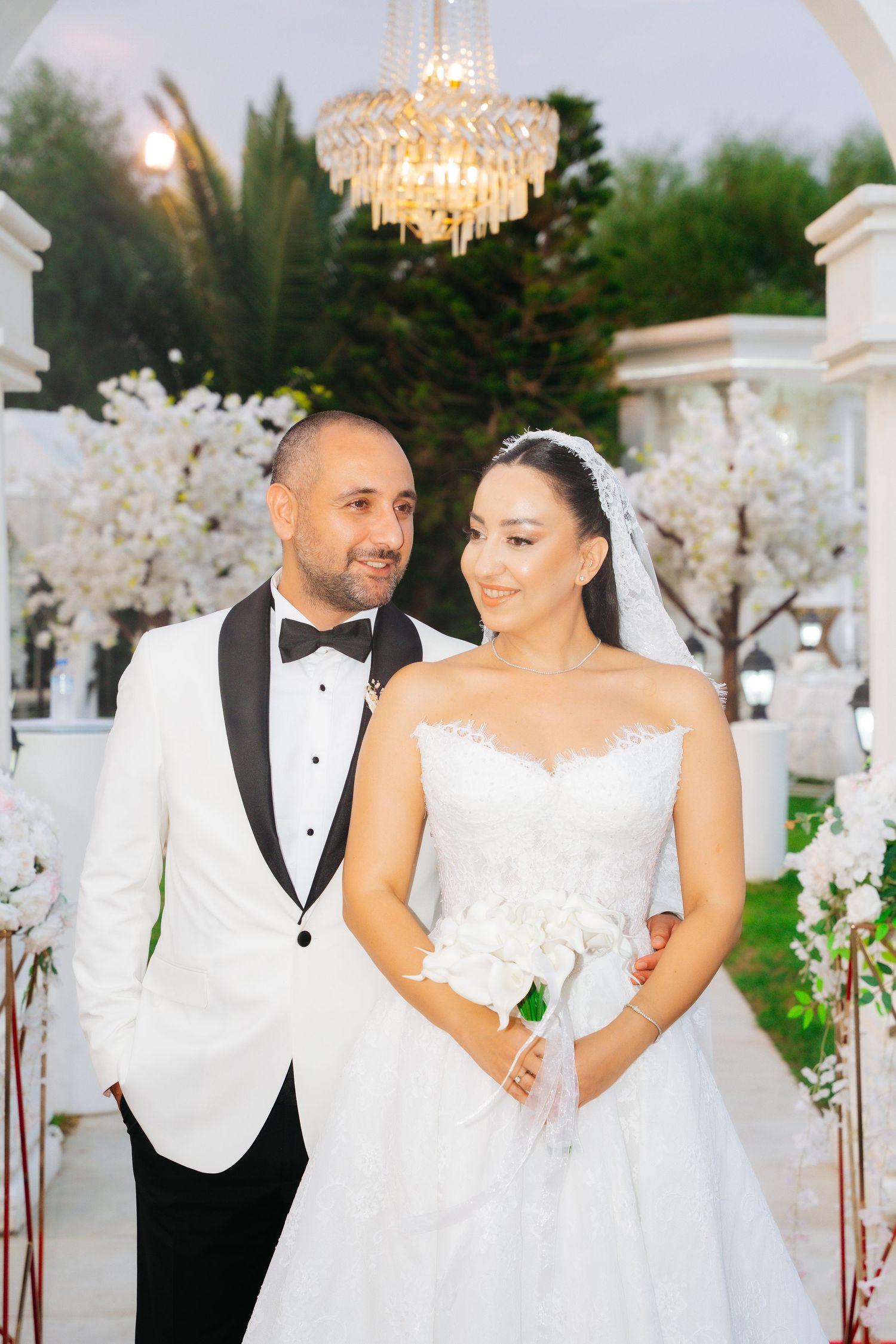 Elegant bride and groom posing at outdoor wedding ceremony with white floral arch and crystal chandelier.