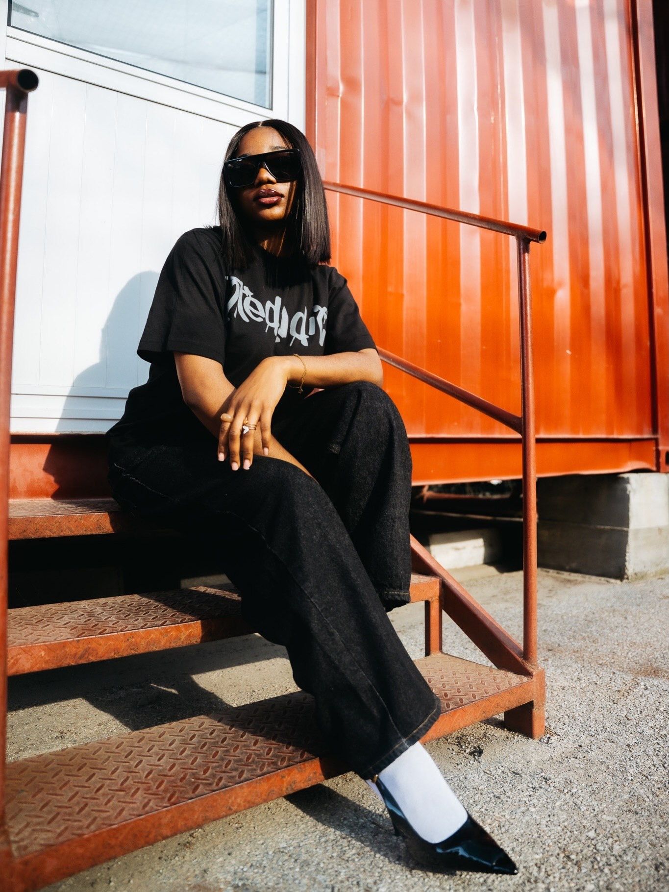 A stylish person in a black graphic tee, wide-leg pants, white socks, and heels sits on metal steps by a red container.