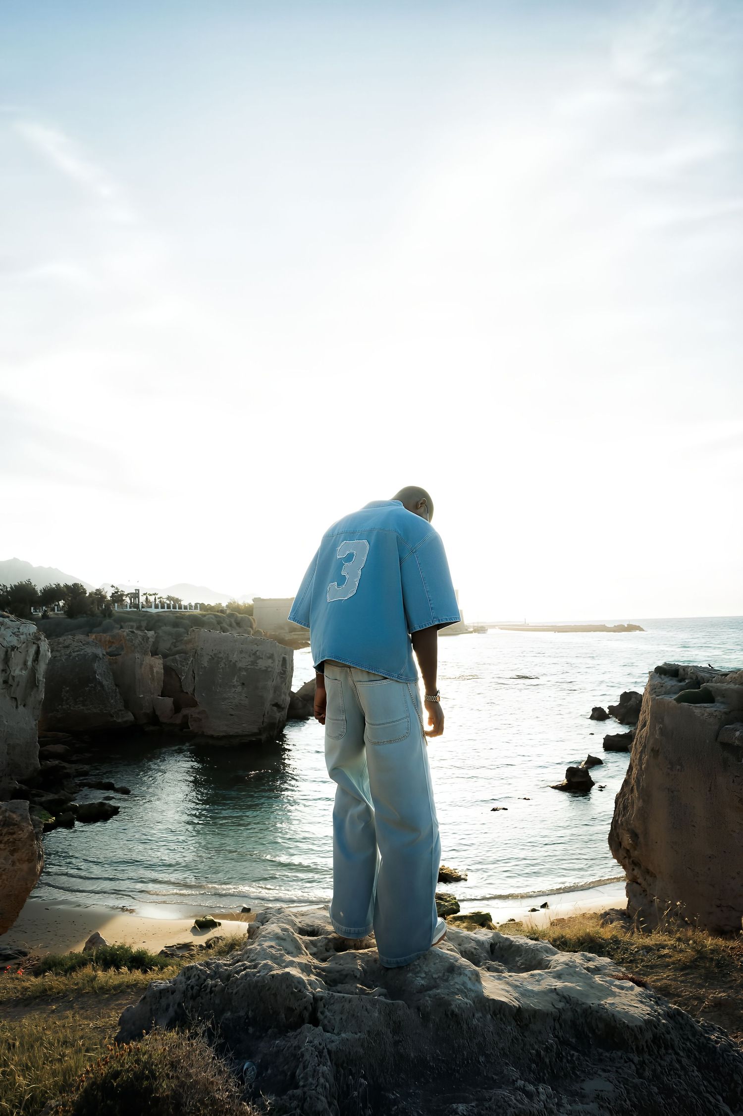 Man in blue number 3 shirt and jeans standing on rocks overlooking a tranquil coastal cove.
