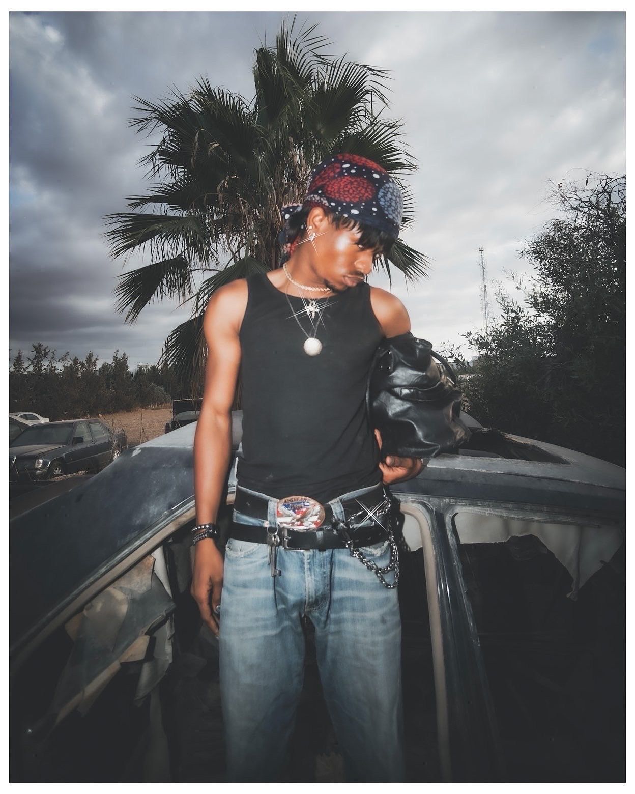 Young man in black tank top, red bandana, jeans, and chain belt poses outdoors near palm tree under cloudy sky.