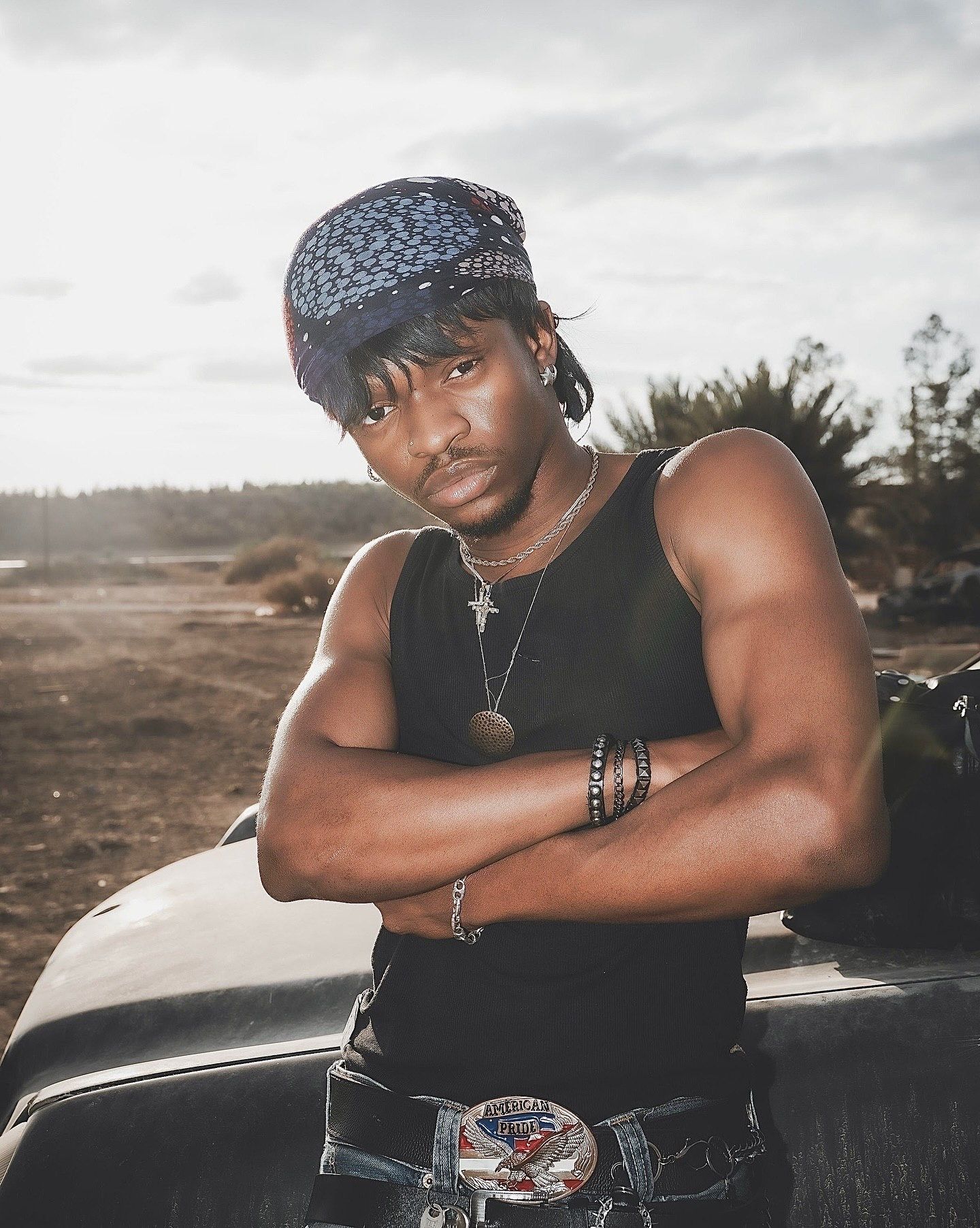 Young man in black tank top, bandana, and cross necklace poses arms crossed against a vehicle outdoors.