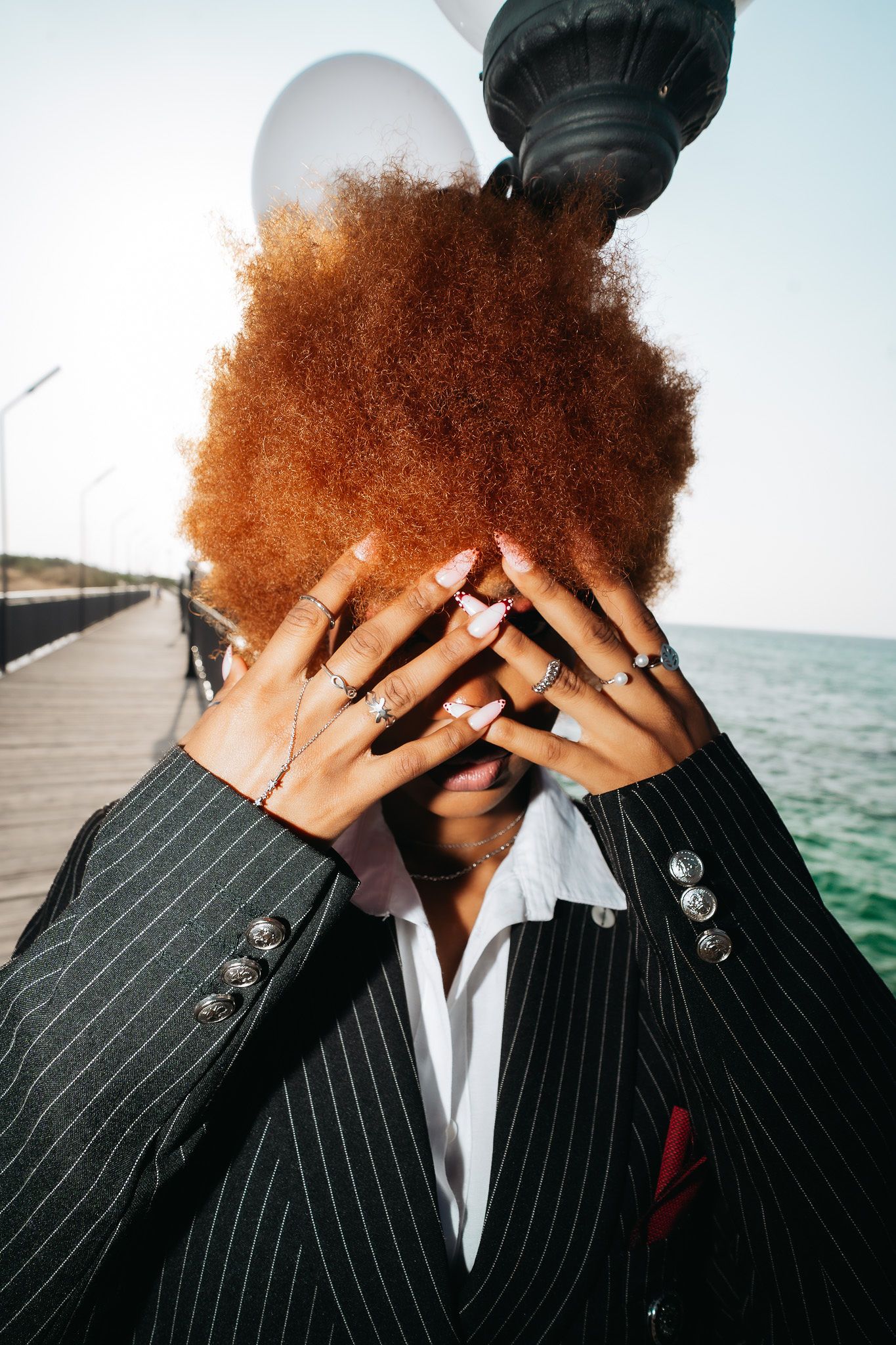 Woman with auburn afro covers face with ringed hands wearing pinstripe blazer by the sea.