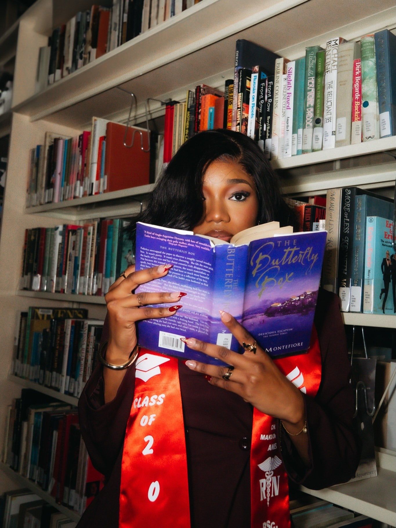 Graduate in red Class sash reads The Butterfly book in front of library bookshelves.