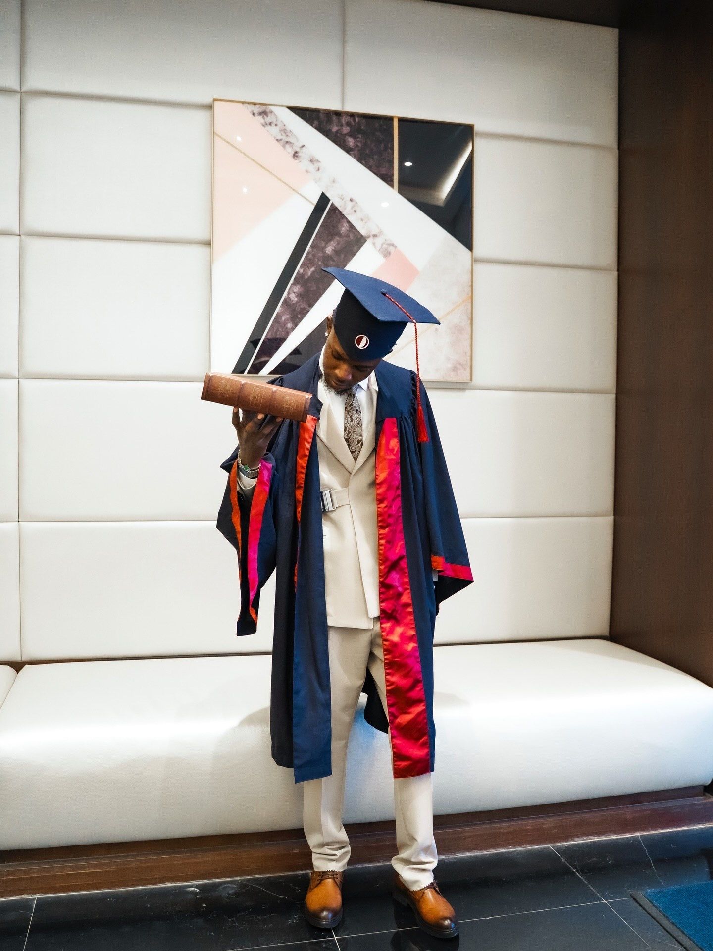 Graduate in navy blue academic gown and cap holds diploma while posing in front of abstract artwork.