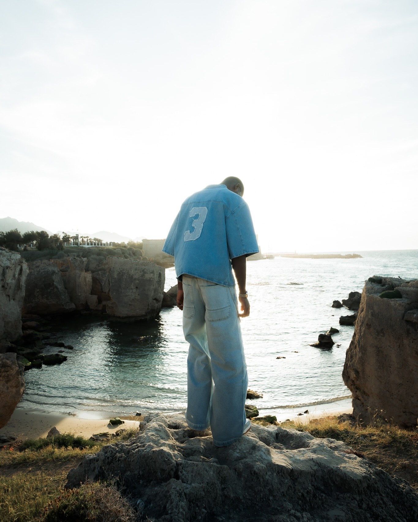 Man in blue jersey and light jeans stands on rocky coastal overlook gazing at sunlit ocean waters.