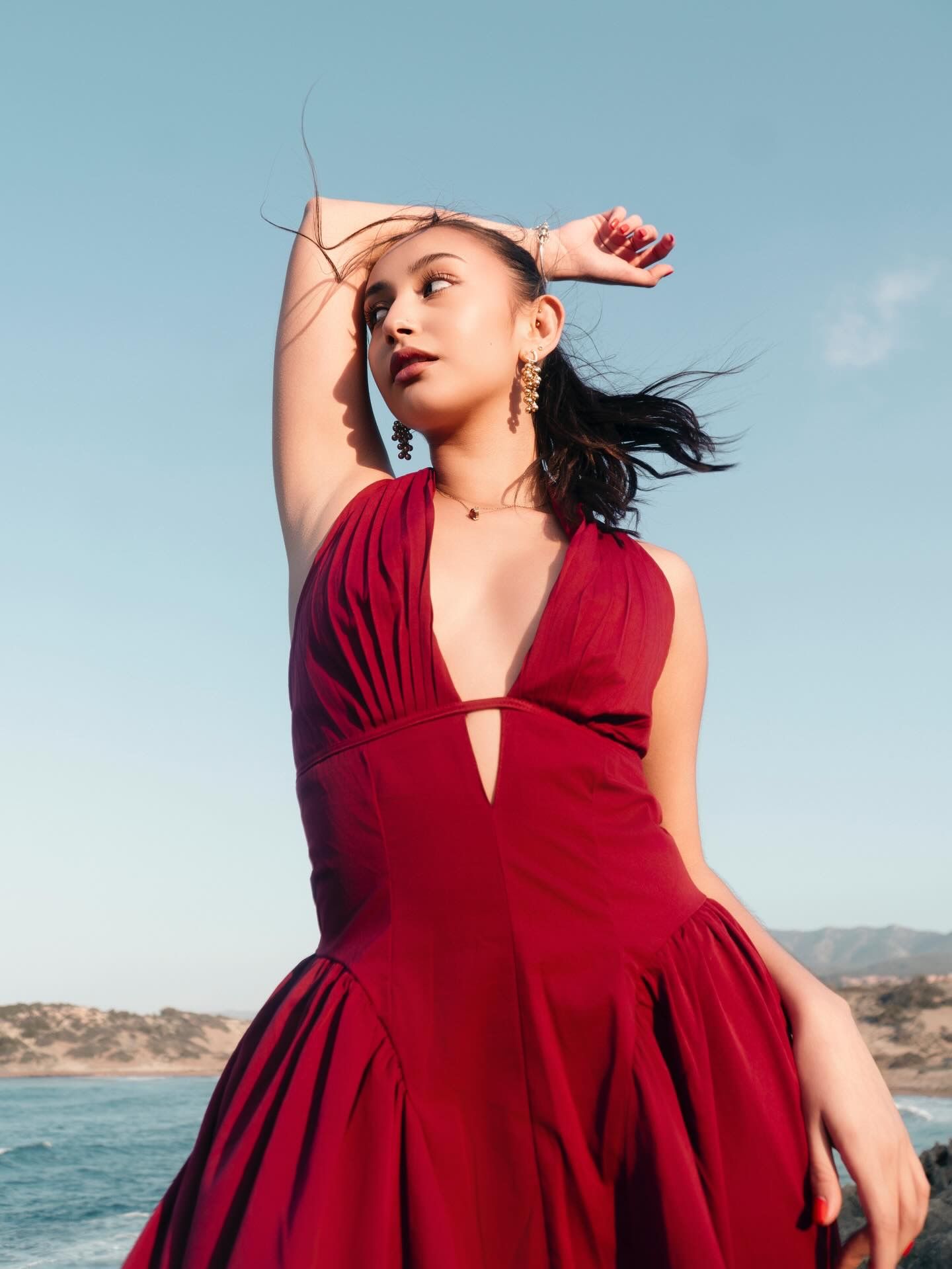 Woman in red halter dress poses on beach with arm raised, ocean and hills in background.