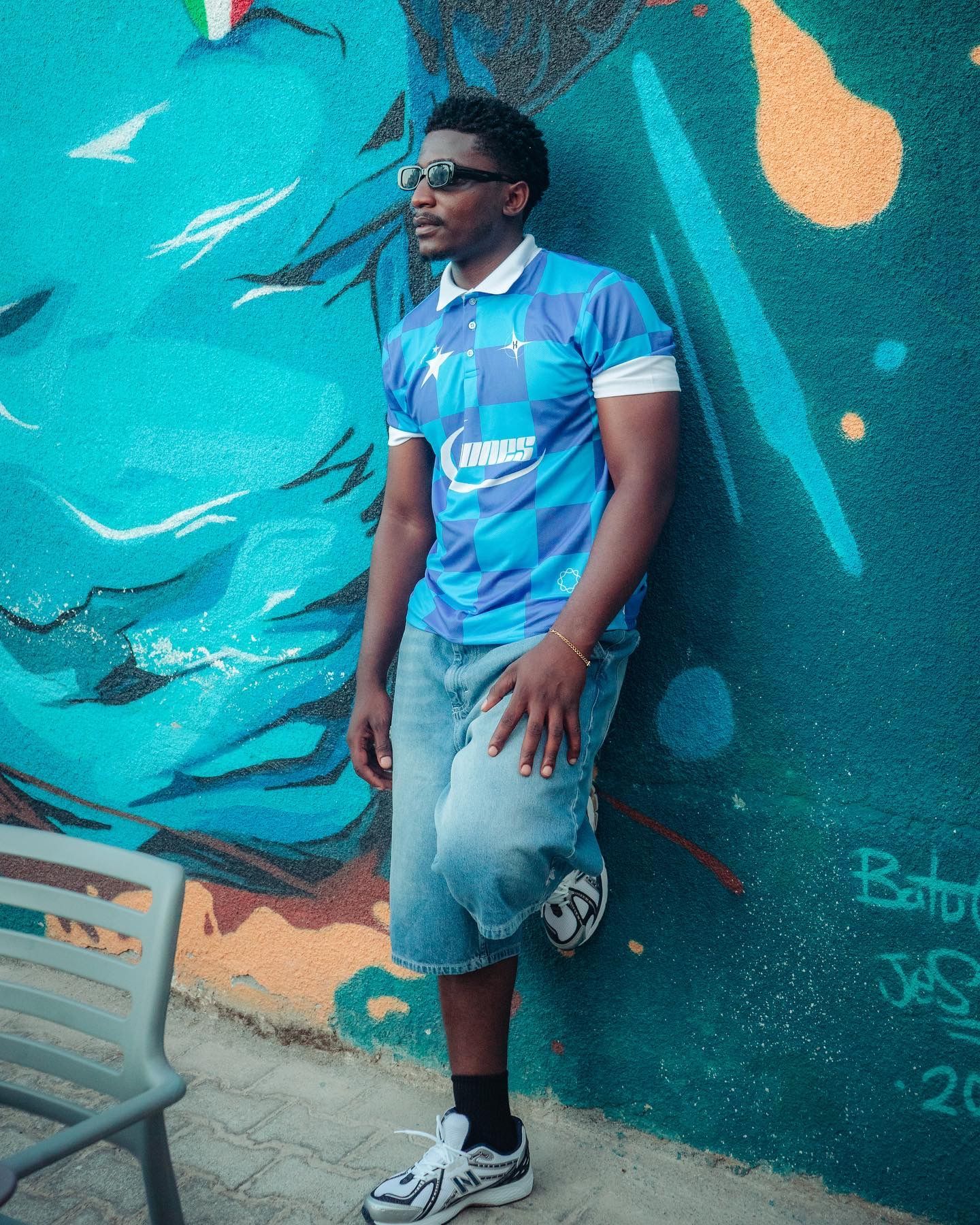 Man in blue soccer jersey and denim shorts posing against colorful graffiti wall wearing sunglasses.