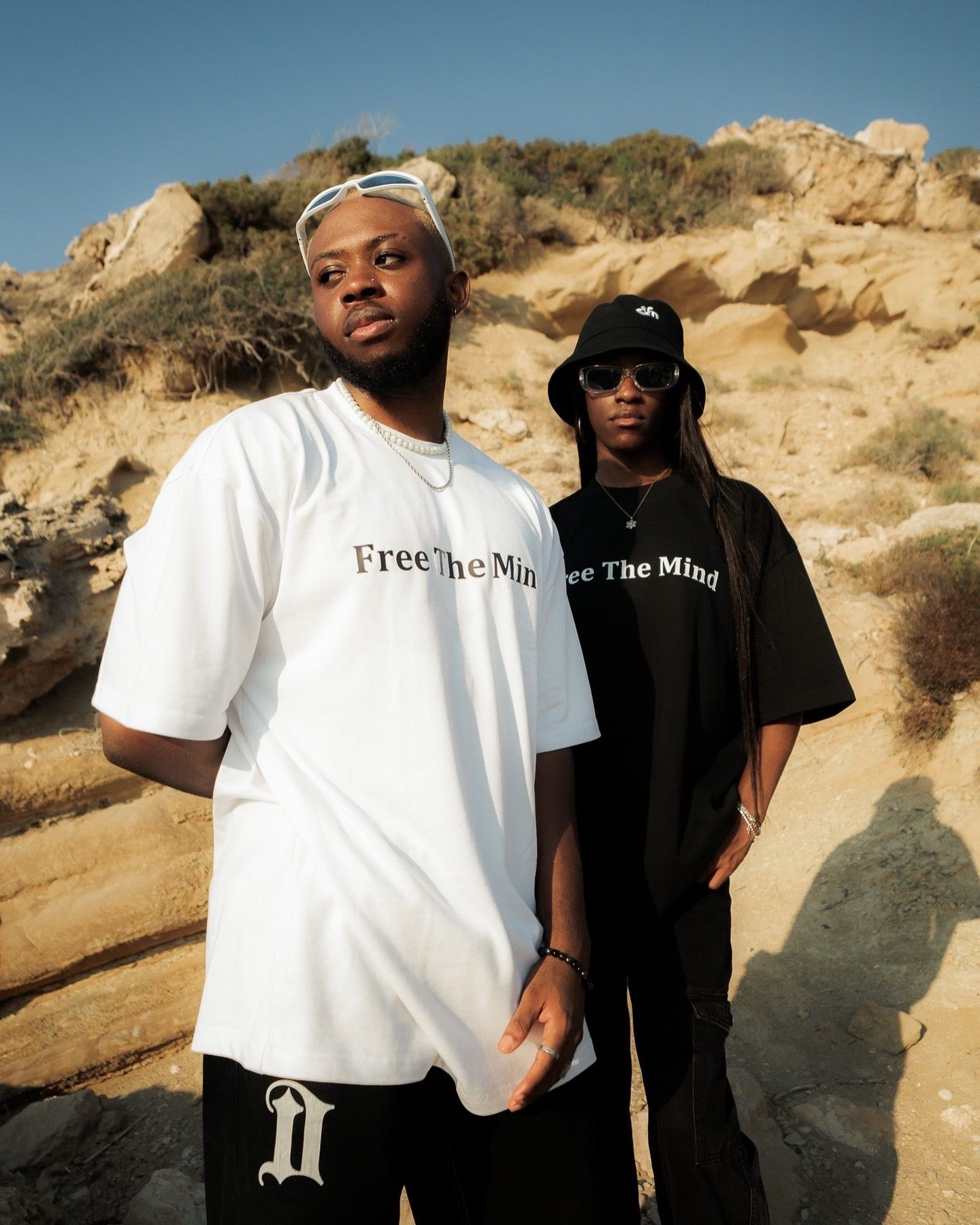 Two men wearing matching Free The Mind graphic tees in white and black, posing outdoors near sandy rock formations.