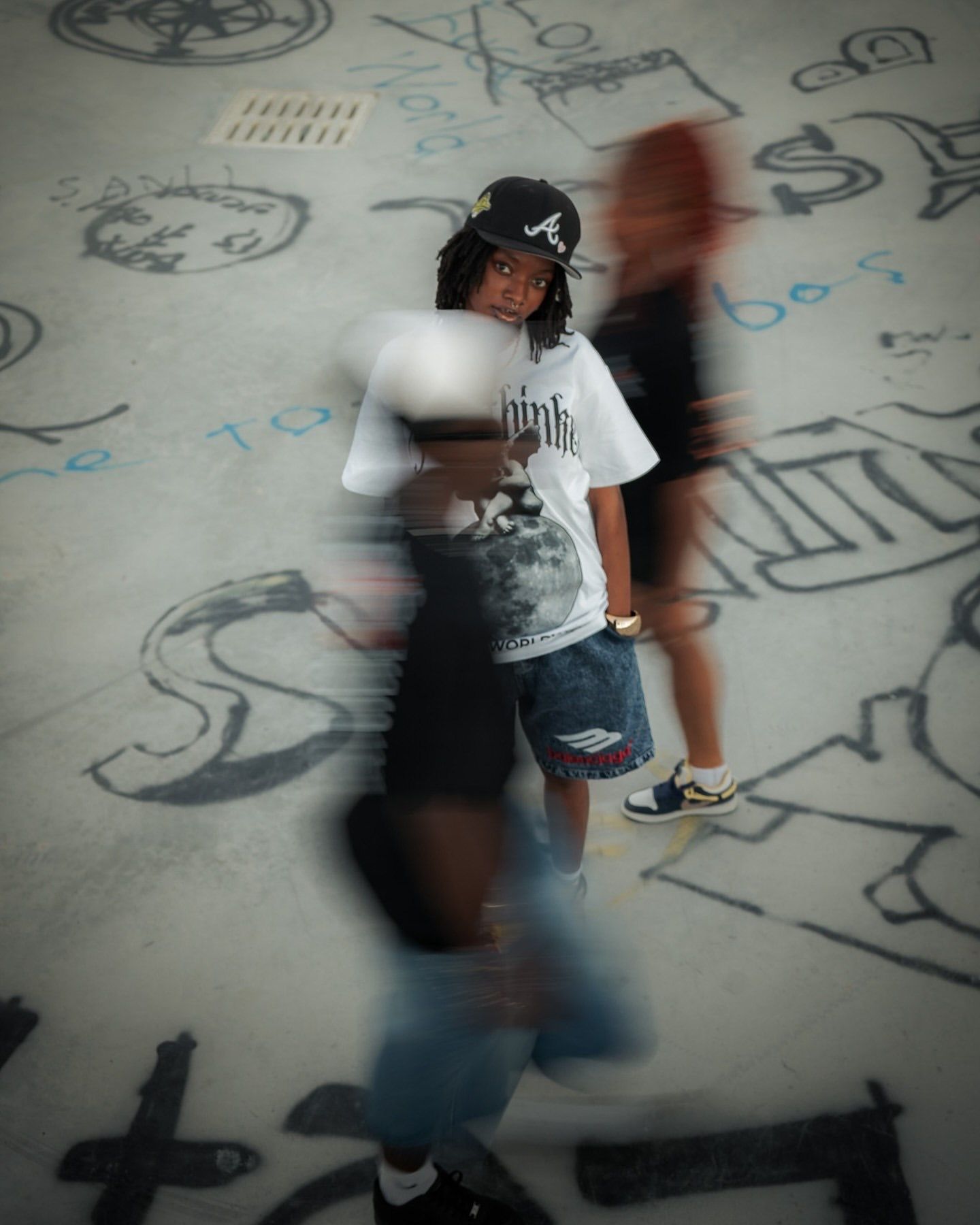 Young skater in white graphic tee and denim shorts poses at skate park with motion-blurred figures and graffiti walls.