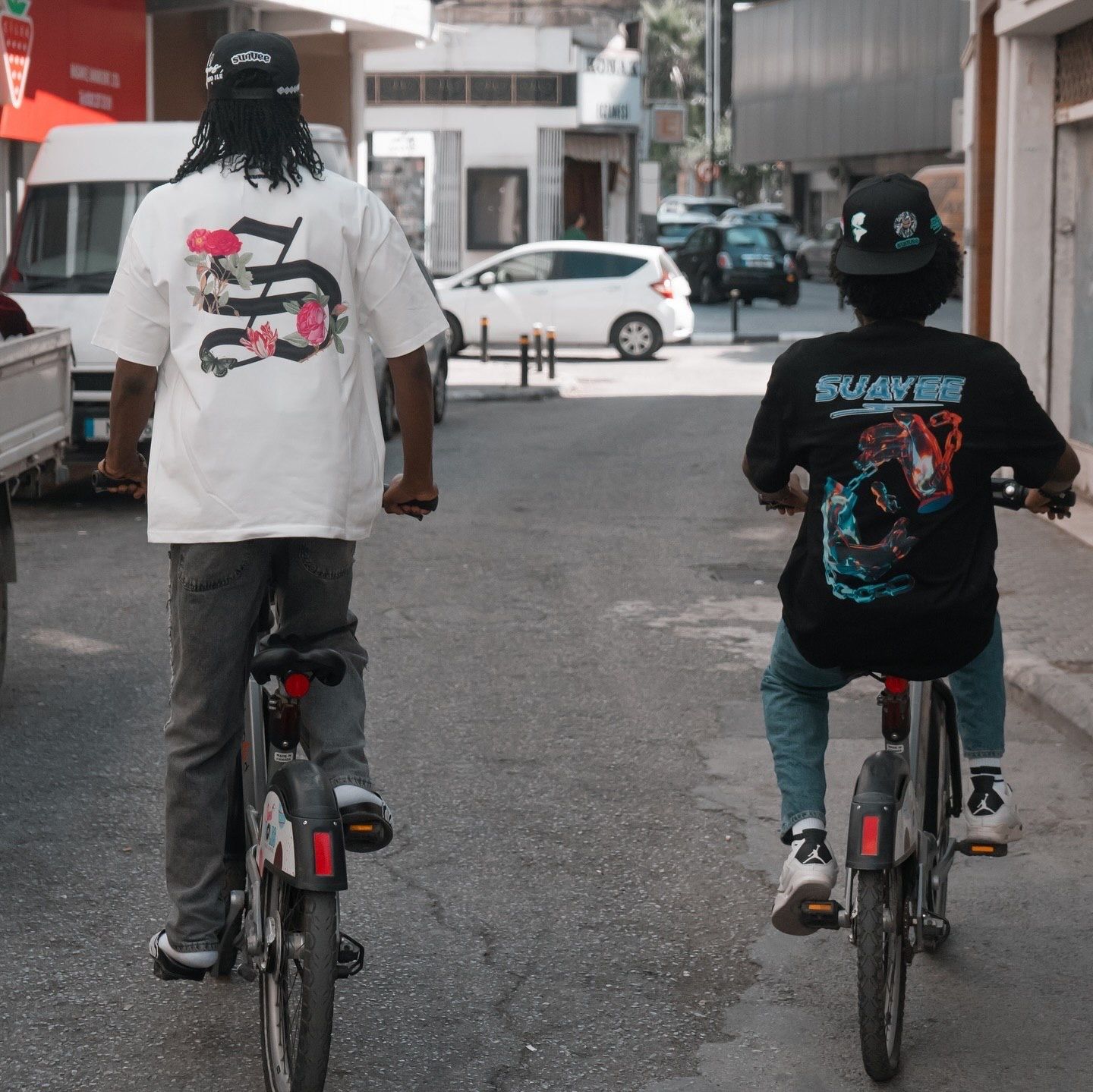 Two people riding bicycles down a urban street, one wearing a white graphic tee and the other a black Spiderman shirt.